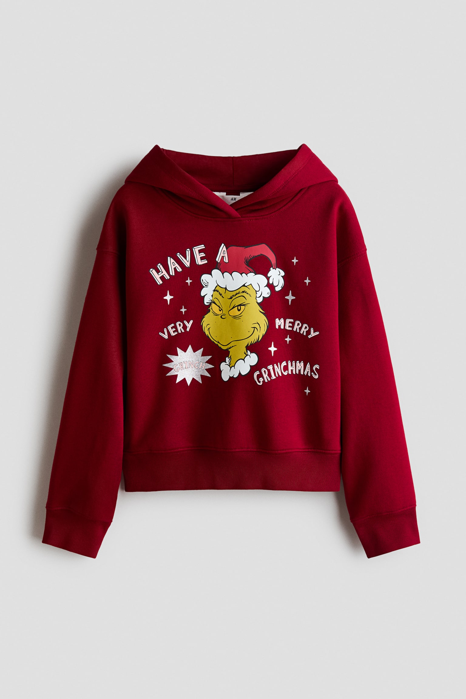 A dark red loose-fit hoodie is shown from the front, featuring a printed graphic of The Grinch wearing a Santa hat and the text "HAVE A VERY MERRY GRINCHMAS" on the chest. The garment displays dropped shoulders, a wrap-front hood, and ribbing at the cuffs and hem.