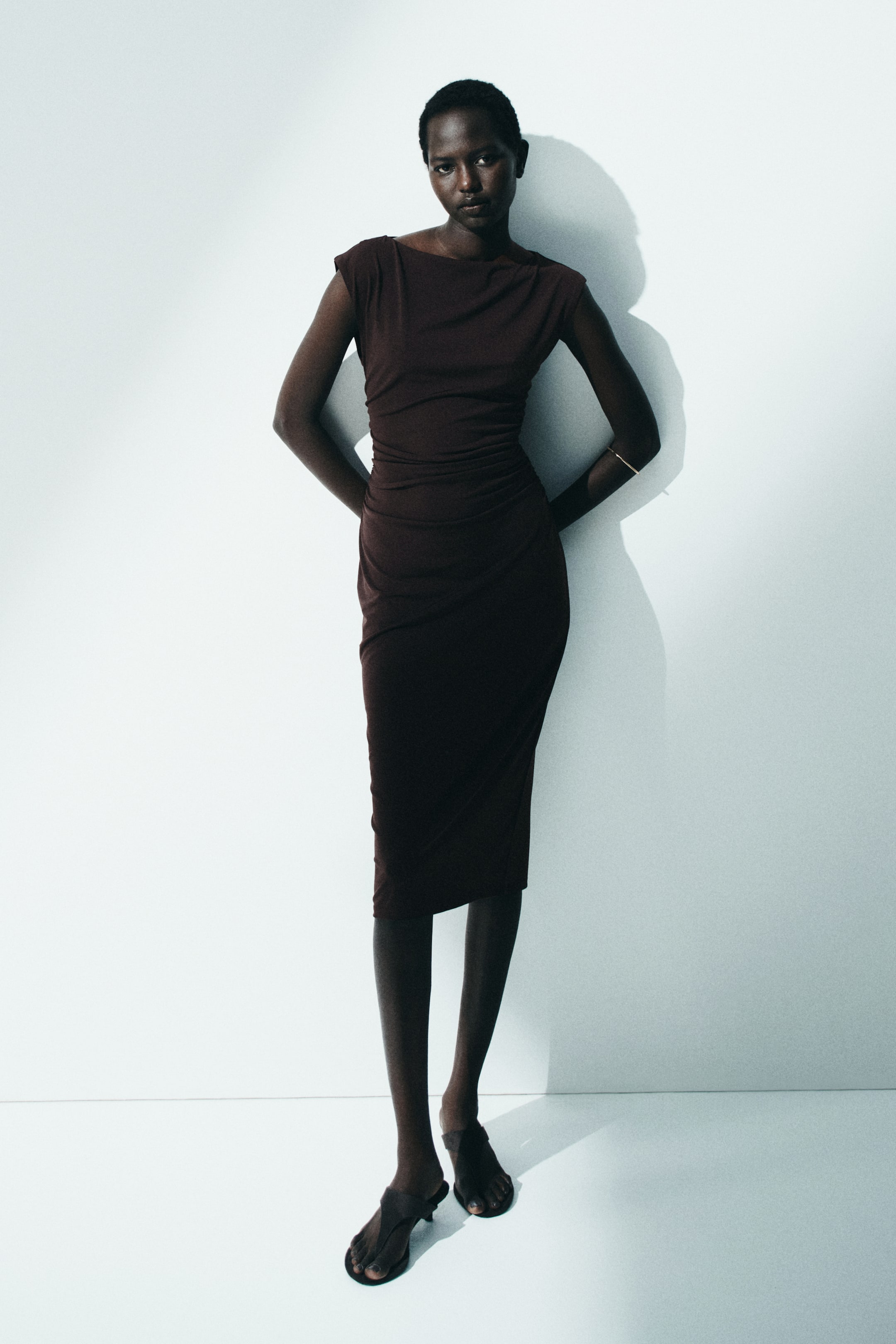 Zobacz większy obraz: Modelka ma na sobie dopasowaną, ciemno brązową sukienkę o długości do łydki wykonaną z elastycznego dżerseju. Sukienka charakteryzuje się dekoltem w łódkę, krótkimi rękawkami i marszczeniami w talii, tworzącymi drapowaną sylwetkę. Modelka ma również na sobie sandały z odkrytymi palcami oraz bransoletkę.