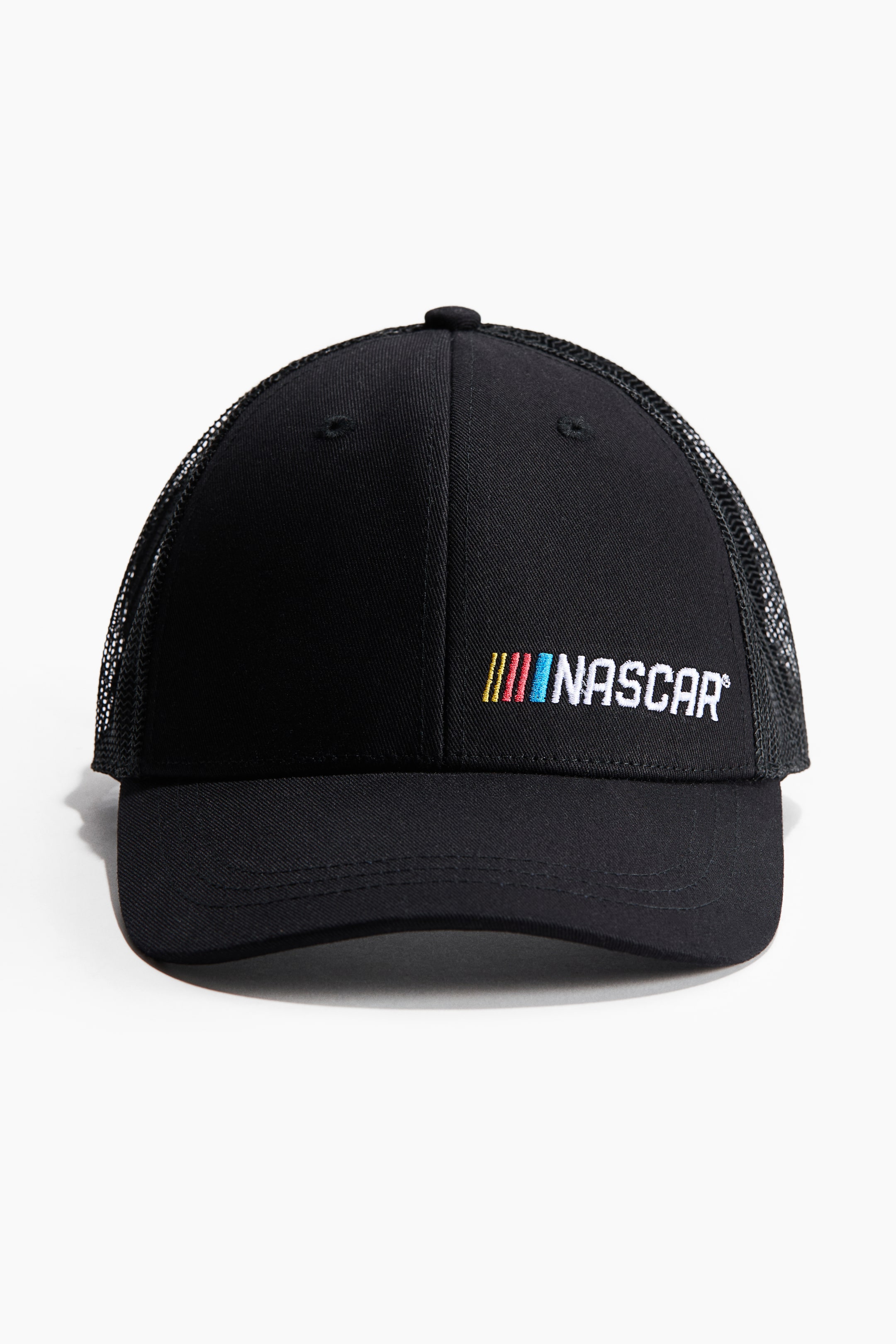 View larger image: A black trucker hat is shown from the front. The solid black front panels feature an embroidered white NASCAR logo to the right of four vertical stripes in yellow, red, light blue, and dark blue. The sides and back are made of black mesh.