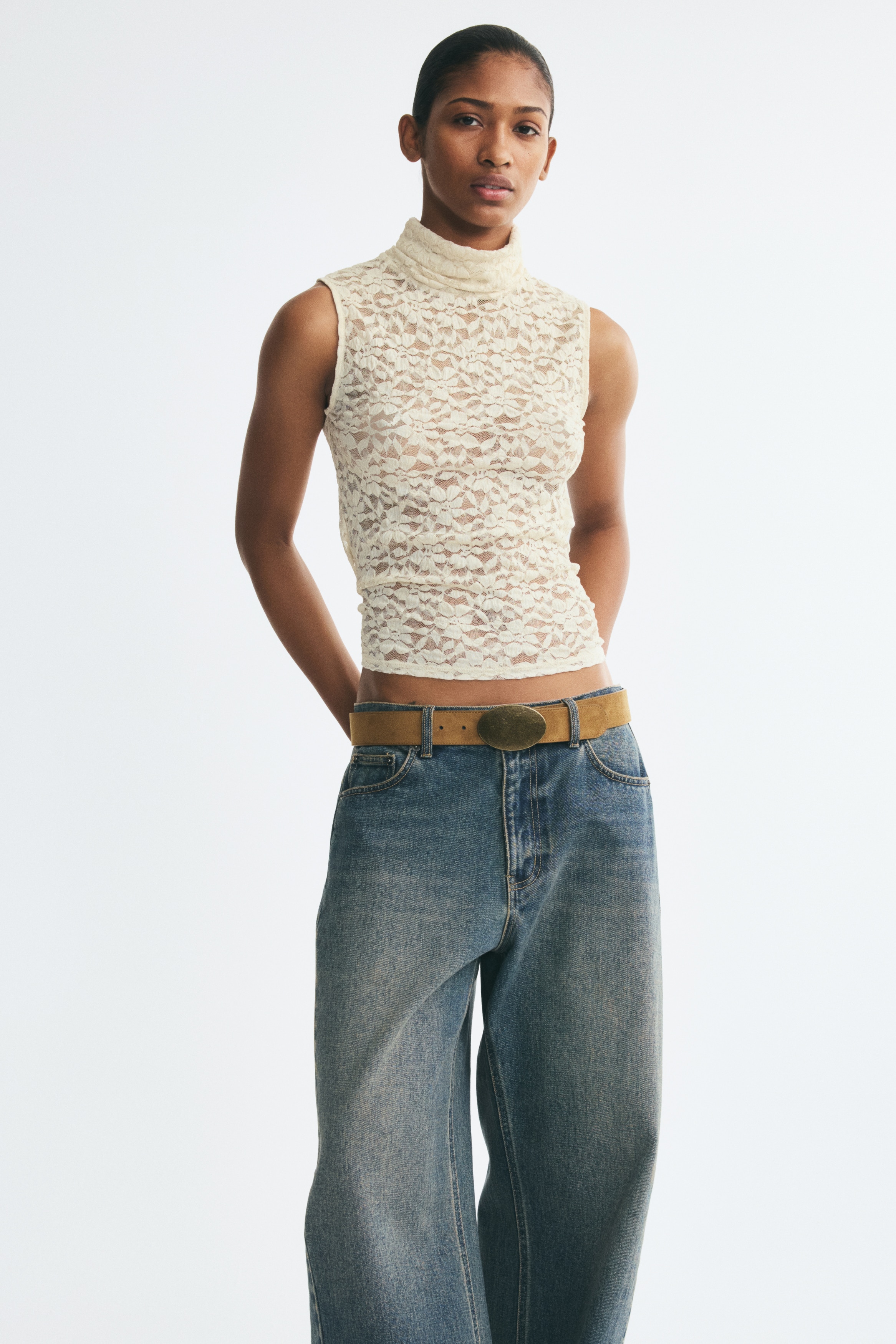 A cream-colored sleeveless lace top is shown from the front, featuring an all-over floral lace pattern and a high, draped turtleneck. The semi-sheer garment is hanging flat, displaying sleeveless armholes and a straight hem.