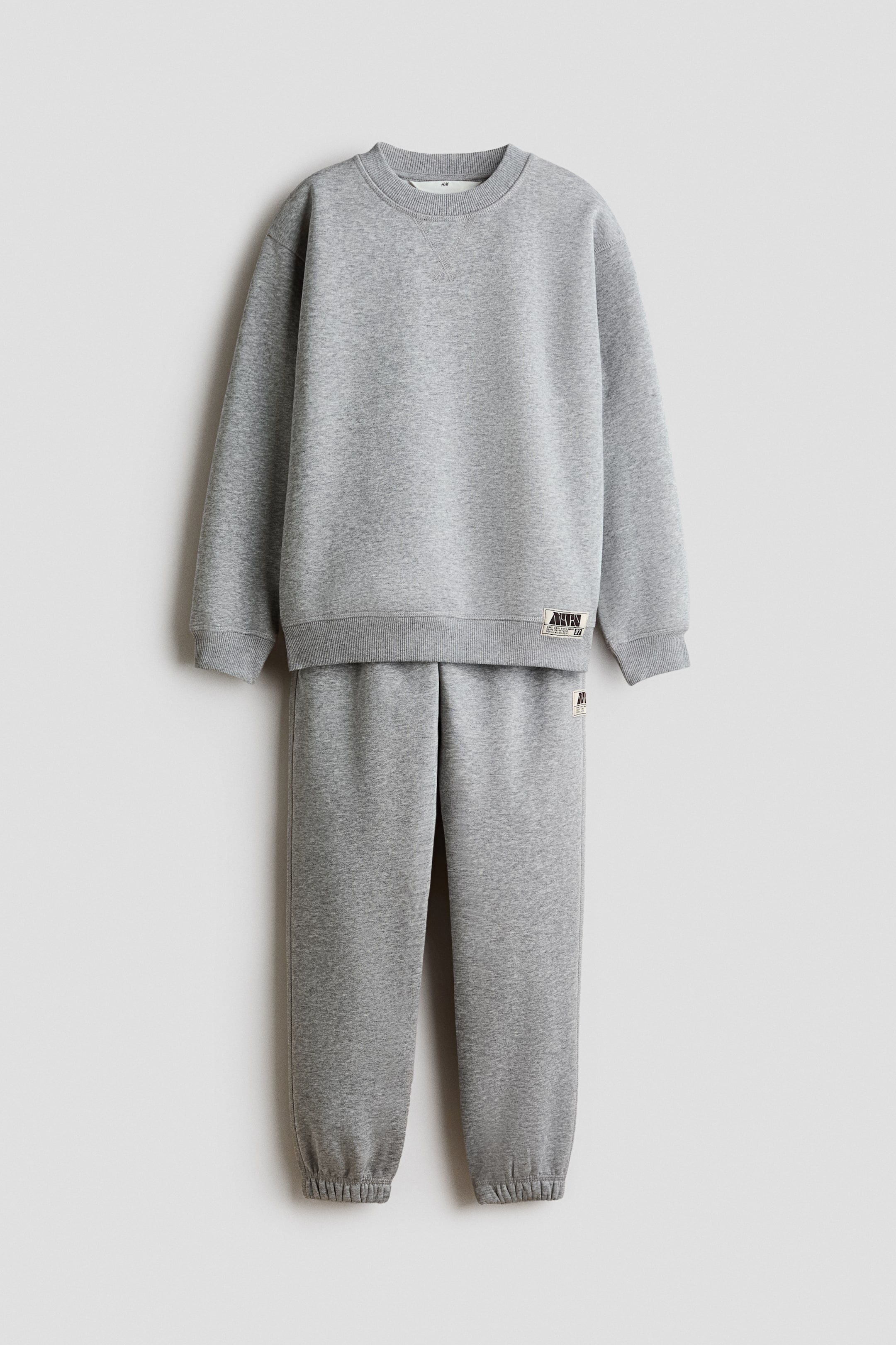 View larger image: A 2-piece brushed-inside sweatshirt set is shown from the front, featuring a grey crew-neck sweatshirt with ribbed collar, cuffs, and hem, along with matching sweatpants with ribbed ankle cuffs. Both pieces feature a small rectangular patch on the front.