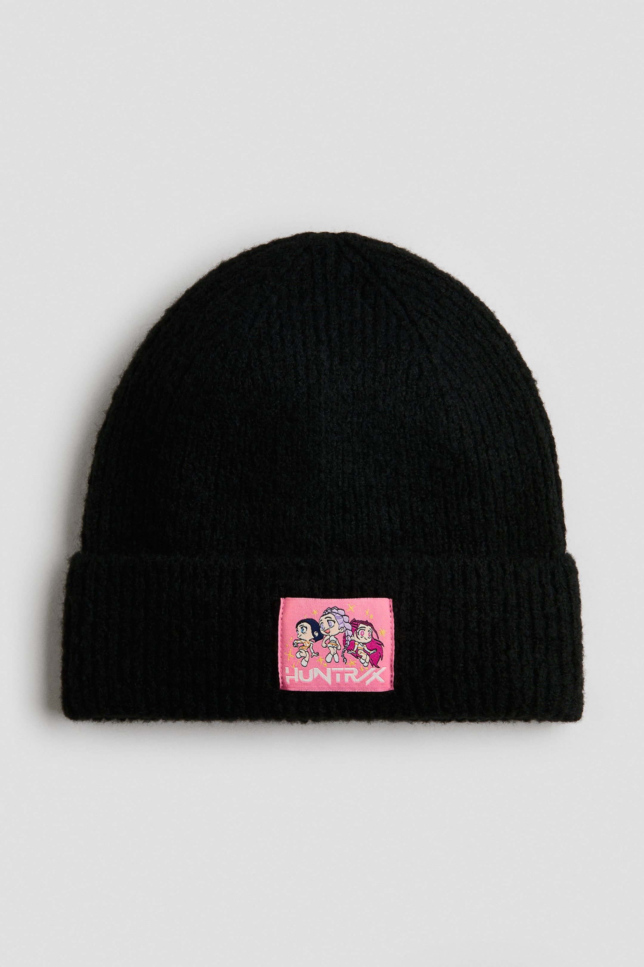 View larger image: A black ribbed-knit beanie is facing forwards, featuring a folded cuff with a rectangular pink patch stitched to the centre. The patch displays three cartoon-style characters above white stylised text reading "HUNTRX".