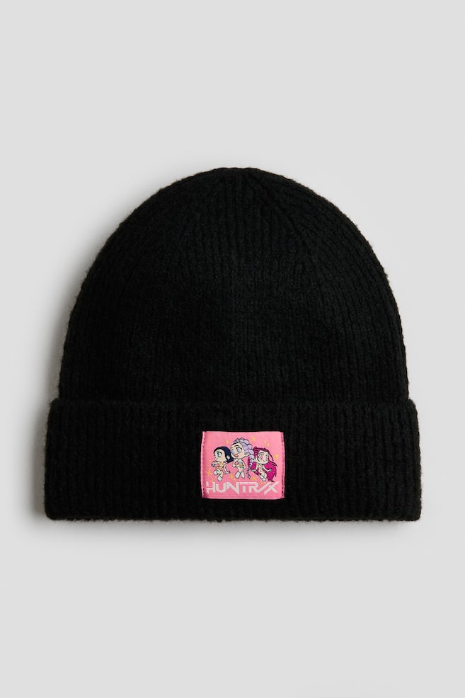 Eine schwarze Beanie aus Rippstrick ist frontal ausgerichtet und verfÃ¼gt Ã¼ber einen umgeschlagenen Saum mit einem rechteckigen, rosa AufnÃ¤her, der mittig aufgenÃ¤ht ist. Auf dem AufnÃ¤her sind drei Figuren im Cartoon-Stil Ã¼ber weiÃer, stilisierter Schrift mit der Aufschrift âHUNTRXâ zu sehen. - 1