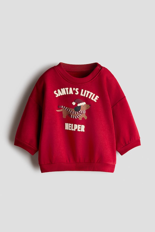 A red print sweater is shown facing forward, featuring a crew neck with a shoulder snap button, ribbed cuffs, and a ribbed hem. The centre displays a graphic of a dachshund wearing a Santa hat and striped scarf, framed by the text "SANTA'S LITTLE HELPER" in white lettering. - 1