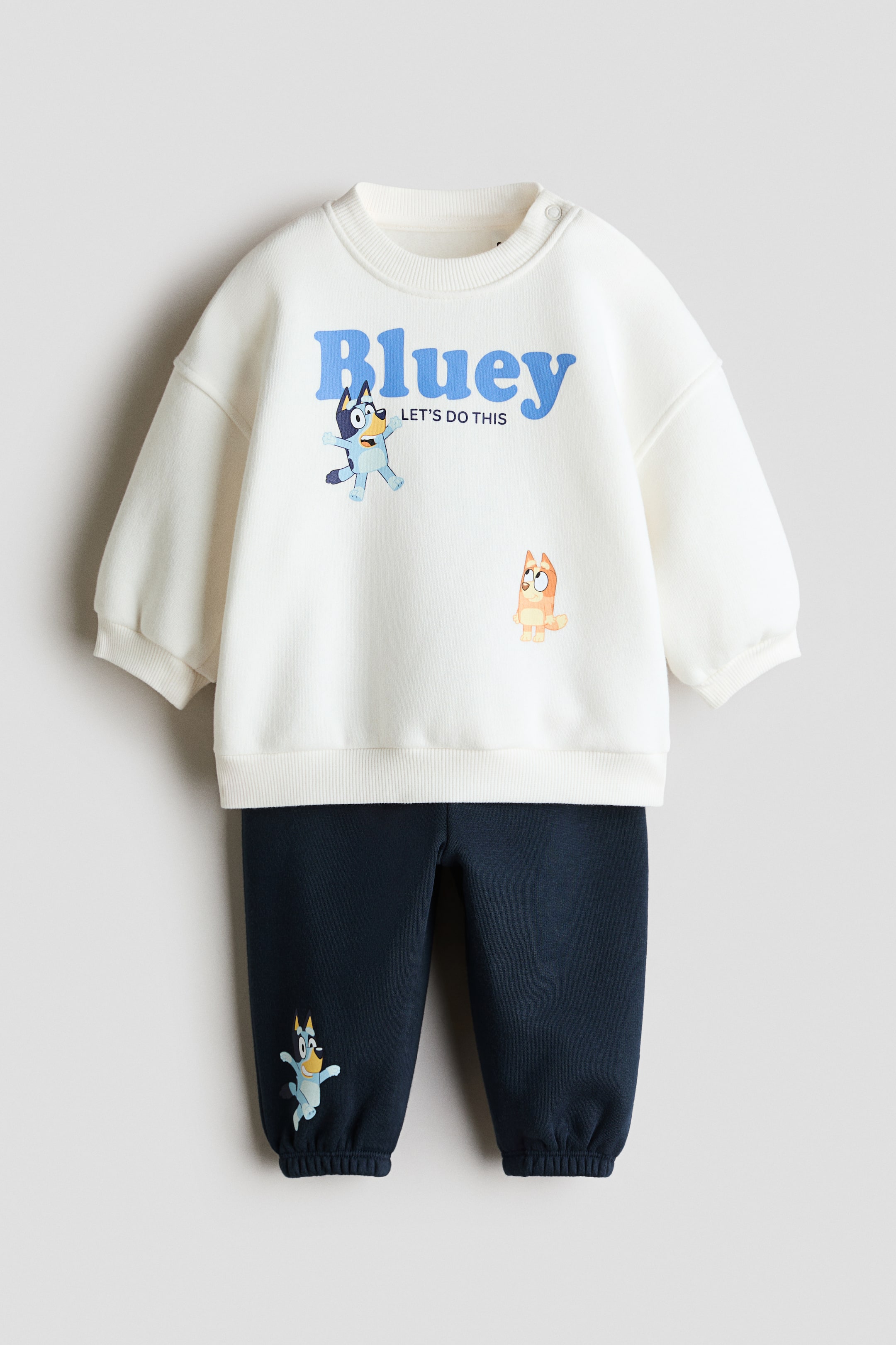 View larger image: This front-facing view displays a two-piece sweat fabric set featuring an off-white sweatshirt and matching navy blue sweatpants. The sweatshirt features a ribbed crew neck with a shoulder snap button, ribbed cuffs, and a "Bluey" graphic print on the front, while the sweatpants feature elasticized cuffs and a small character print on the lower leg.