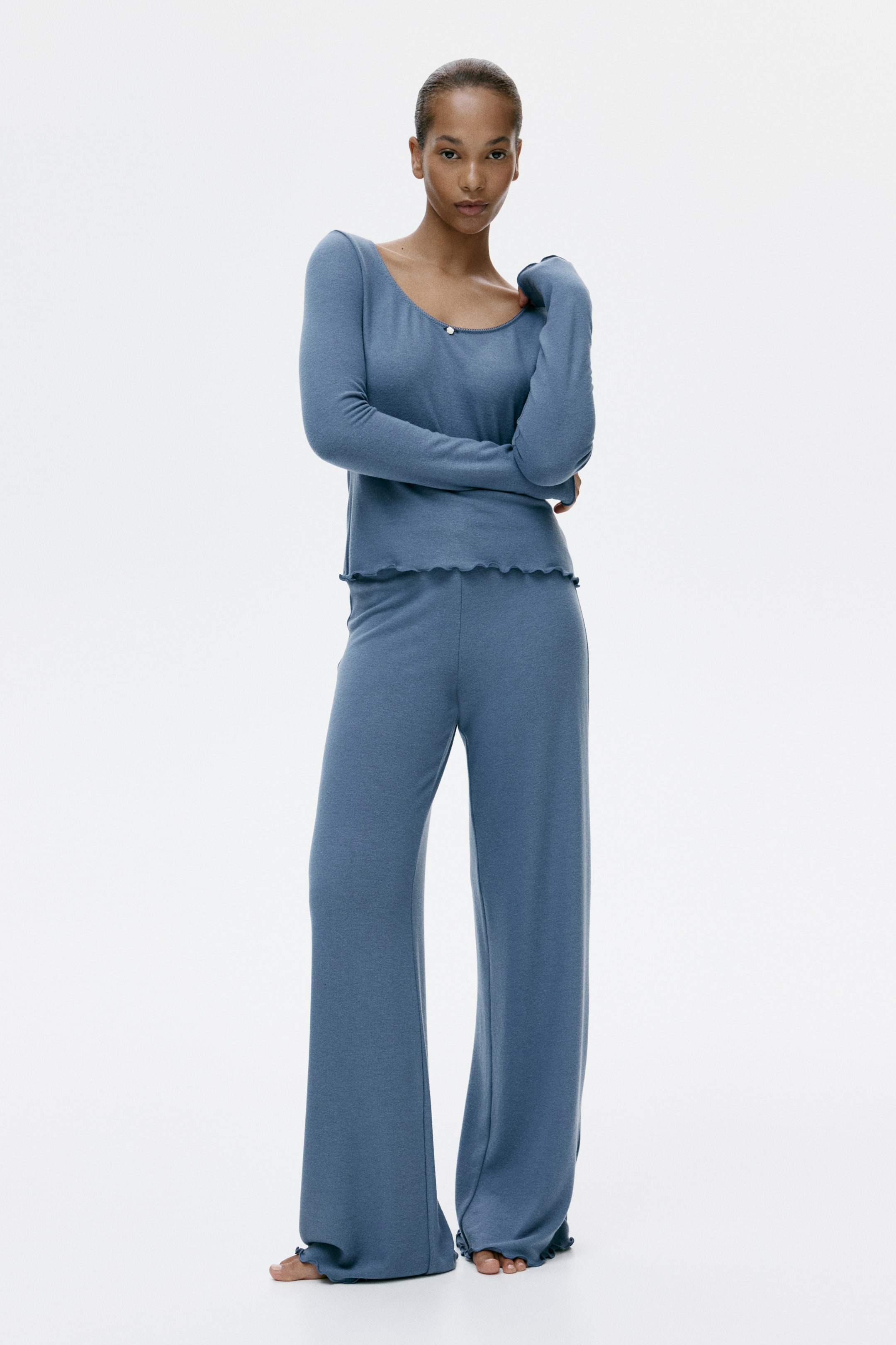 View larger image: A front-facing blue knit loungewear set features a long-sleeved top with a scoop neckline, a single white button detail and a ruffled hem. Matching wide-leg trousers complete the set, also featuring ruffled hems at the ankles.