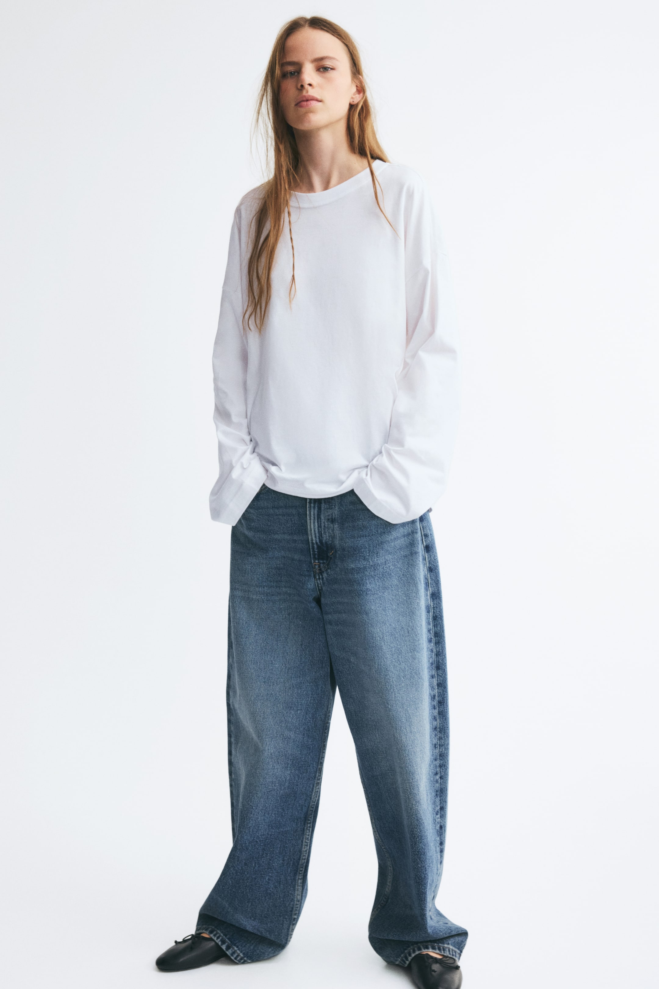 View larger image: A white, loose-fit cotton jersey top featuring a round, rib-trimmed neckline, dropped shoulders, and long sleeves is being worn by the model, who is posing with hands tucked into the pockets of wide-leg blue jeans.