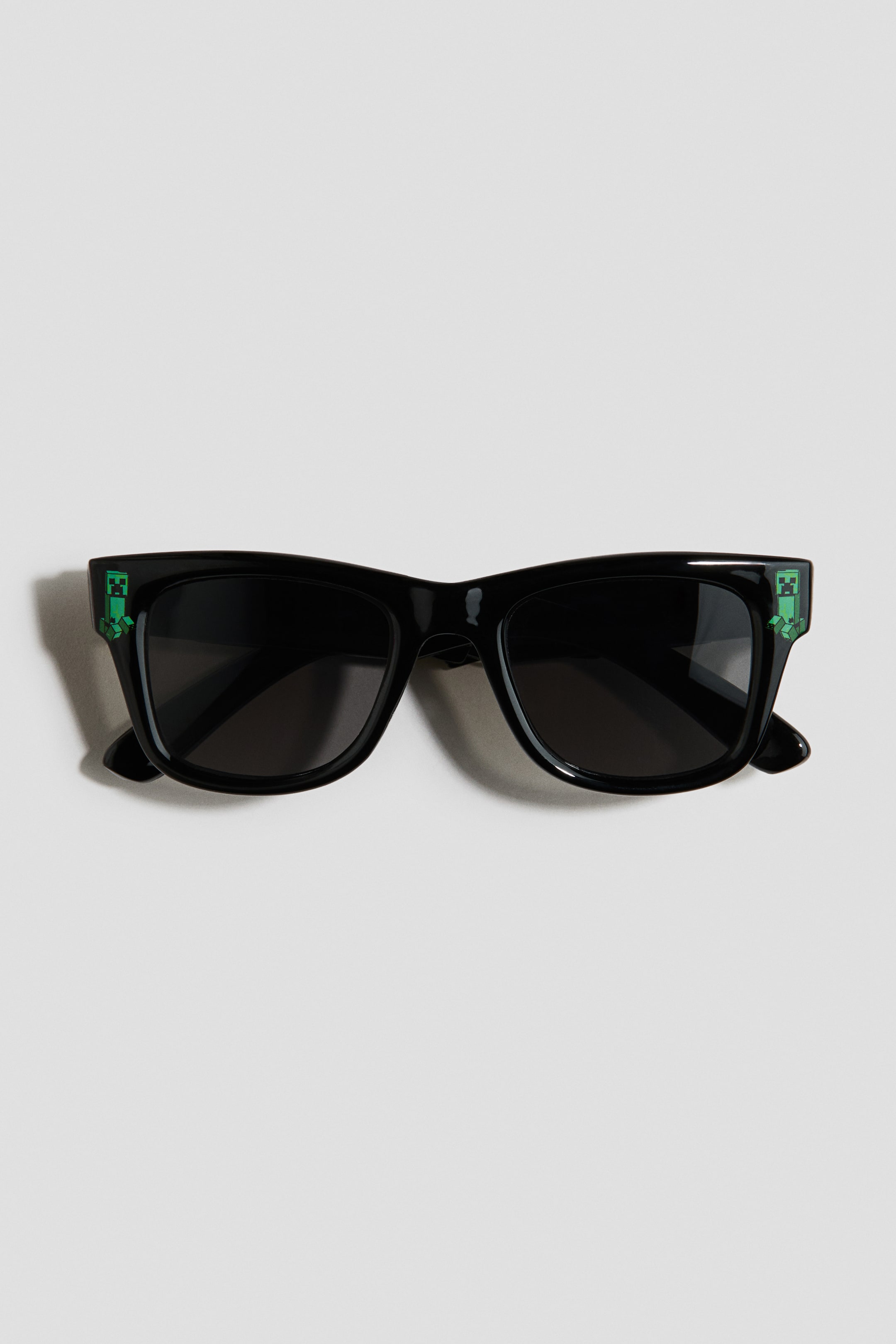 View larger image: The sunglasses feature a black frame with dark lenses, viewed from the FRONT. A green pixelated character is positioned on the outer corner of each frame.