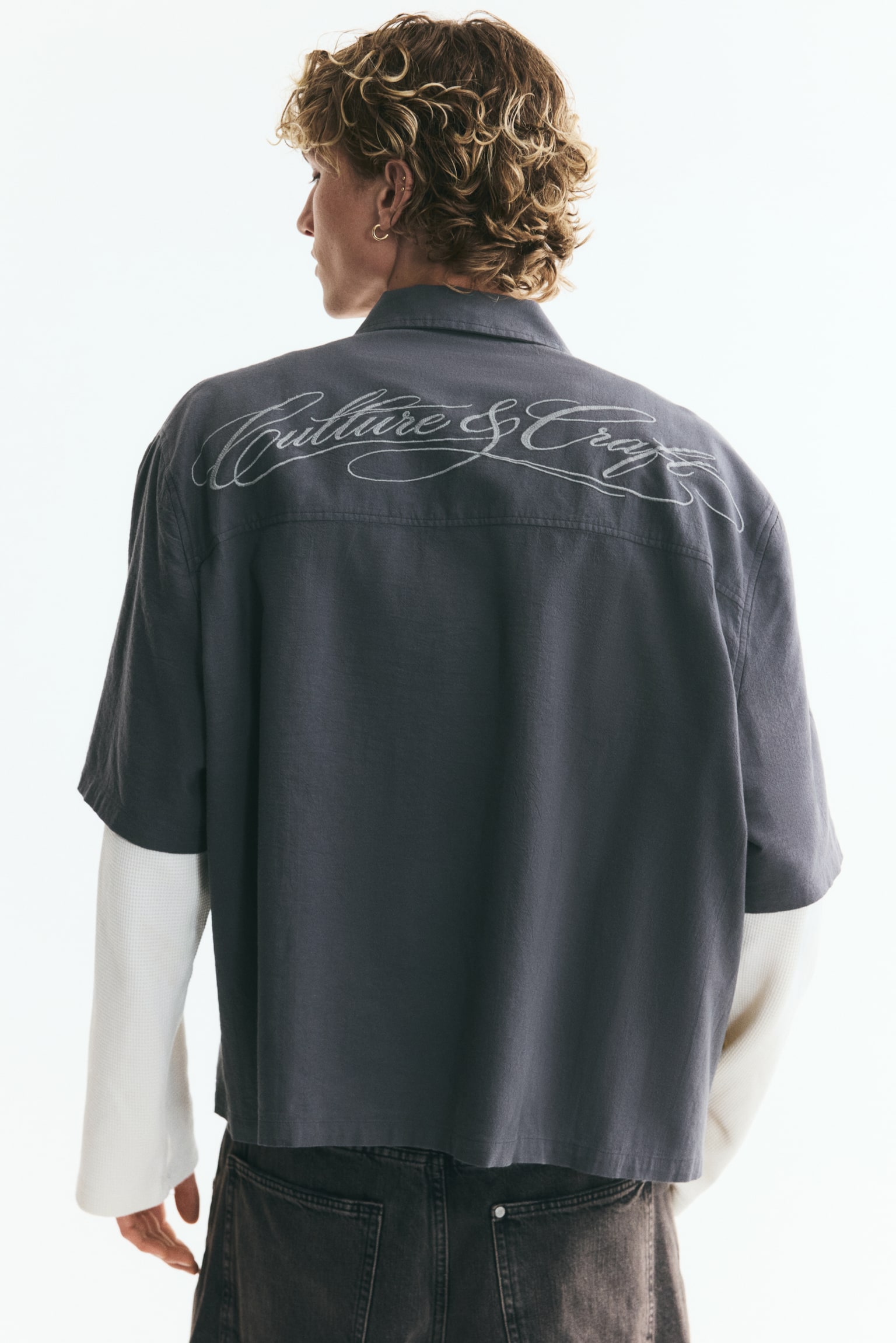 A dark grey short-sleeved collared shirt is layered over a cream-coloured long-sleeved garment. The grey shirt features a full button-down front with dark buttons, whilst the cream sleeves showcase a textured knit fabric. - 1