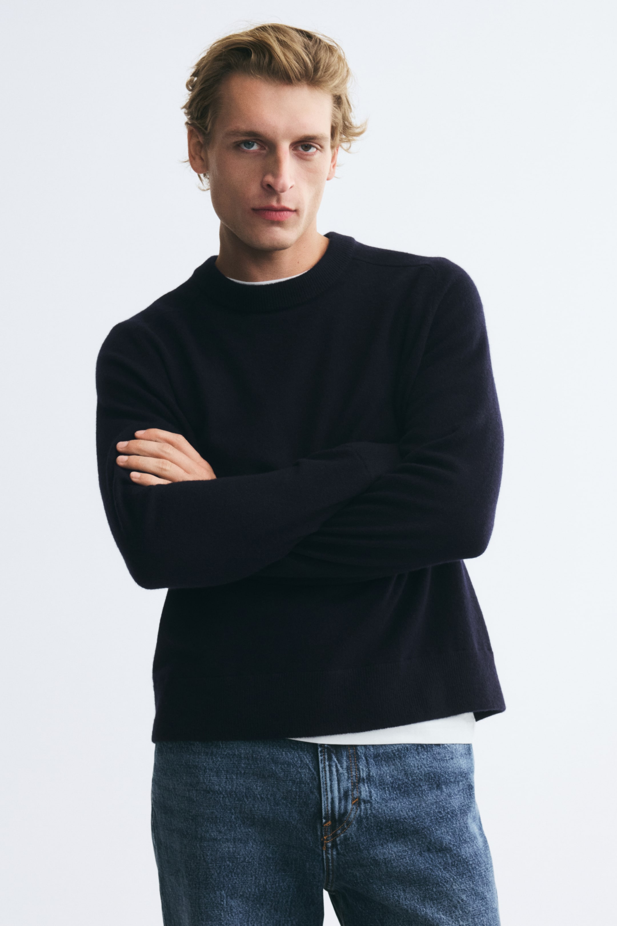 Agrandir l'image: Le mannequin porte un pull col rond bleu marine foncé, debout les bras croisés. Ce pull en maille présente un col, des poignets et un ourlet côtelés, et est porté sur un t-shirt blanc avec un jean bleu.