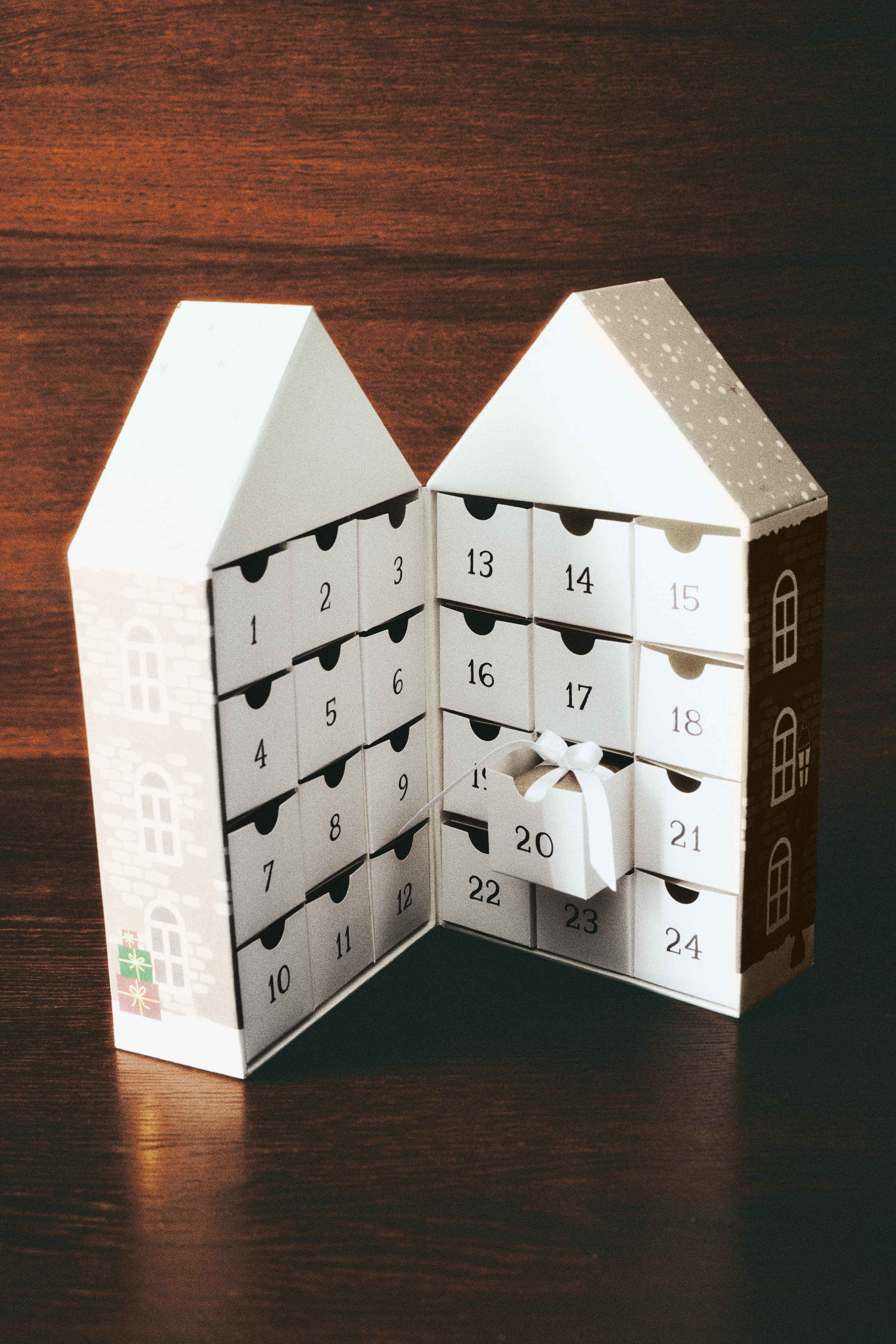 Een vooraanzicht van een huisvormige geschenkdoos toont een bruin baksteenontwerp met witte gebogen ramen, een rode dubbele deur en twee groene kerstbomen. De doos is ook versierd met groene slingers en kransen, geaccentueerd met rode strikken.