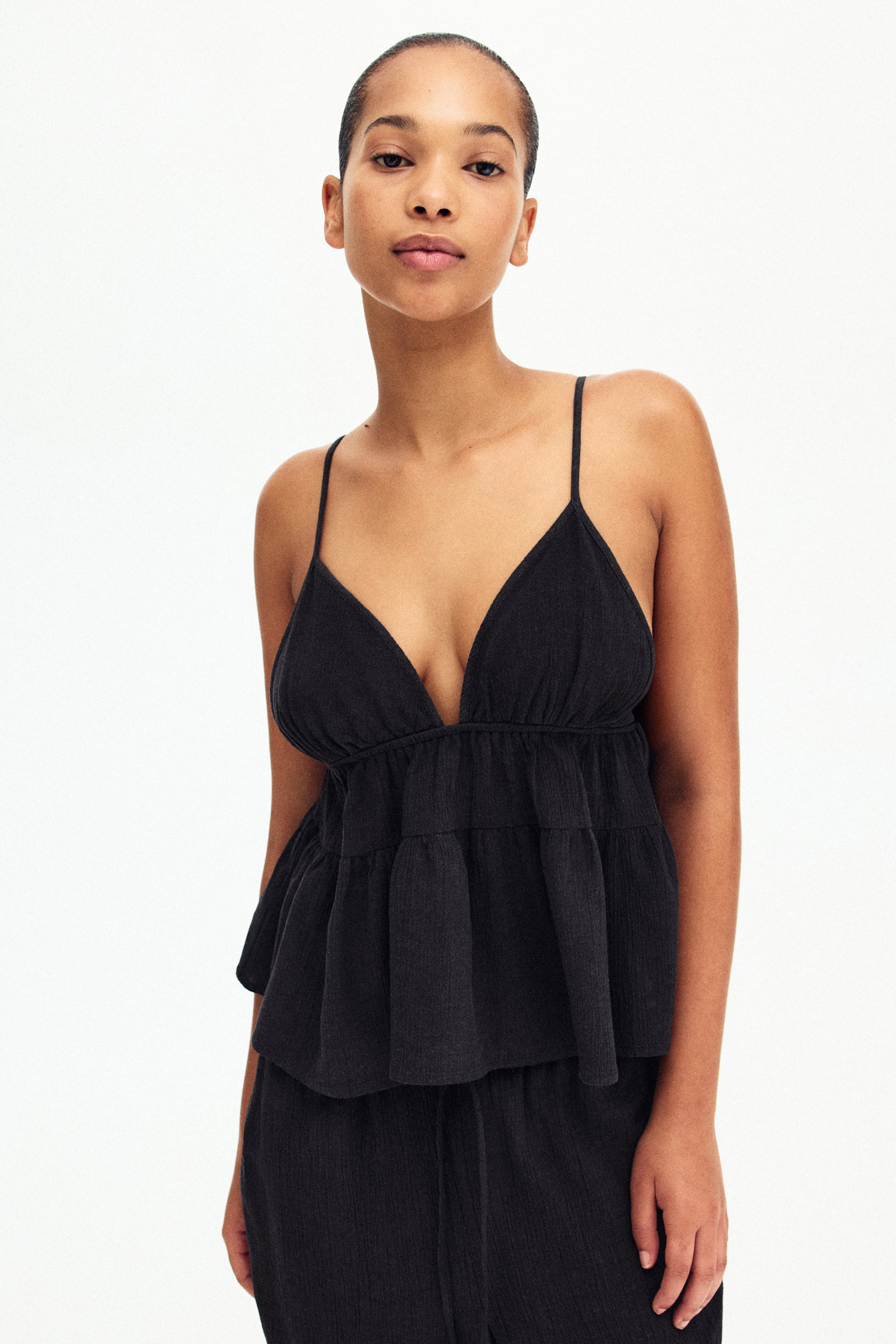 View larger image: The model is donning a black top crafted from a crinkled linen blend weave. It features a V-neckline and narrow adjustable shoulder straps, complemented by gathered elastication under the bust. A gathered seam sits above the waist, leading to a gently flared hem. The ensemble is completed with matching crinkled bottoms.