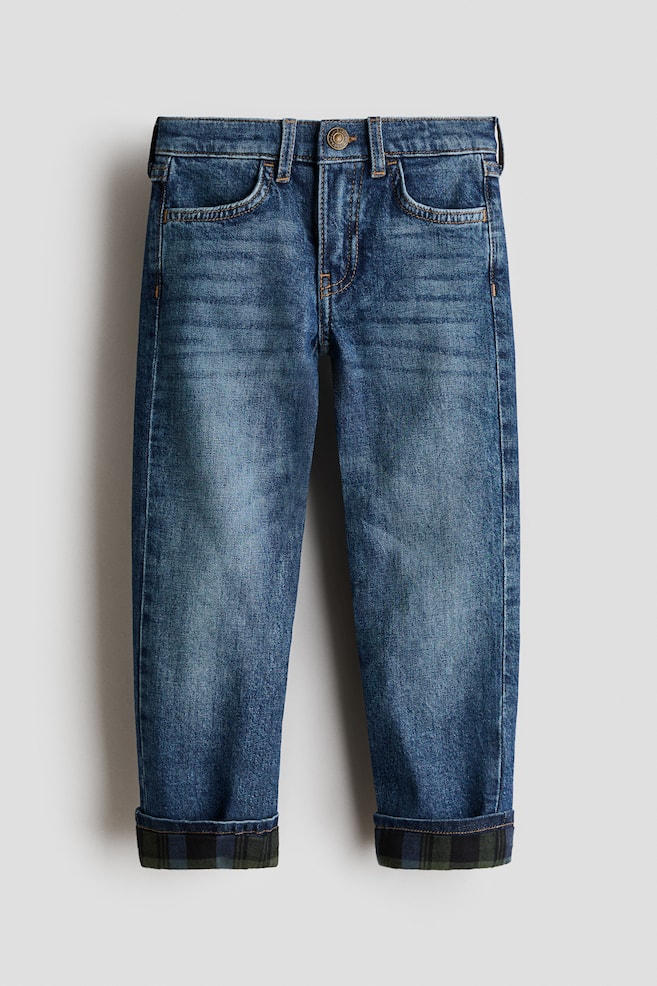 A pair of blue washed denim jeans is shown from the front, featuring a classic five-pocket design, belt loops and a central button fastening at the waist. The straight-leg jeans have turned-up cuffs, revealing a dark green and blue plaid fabric lining. - 1