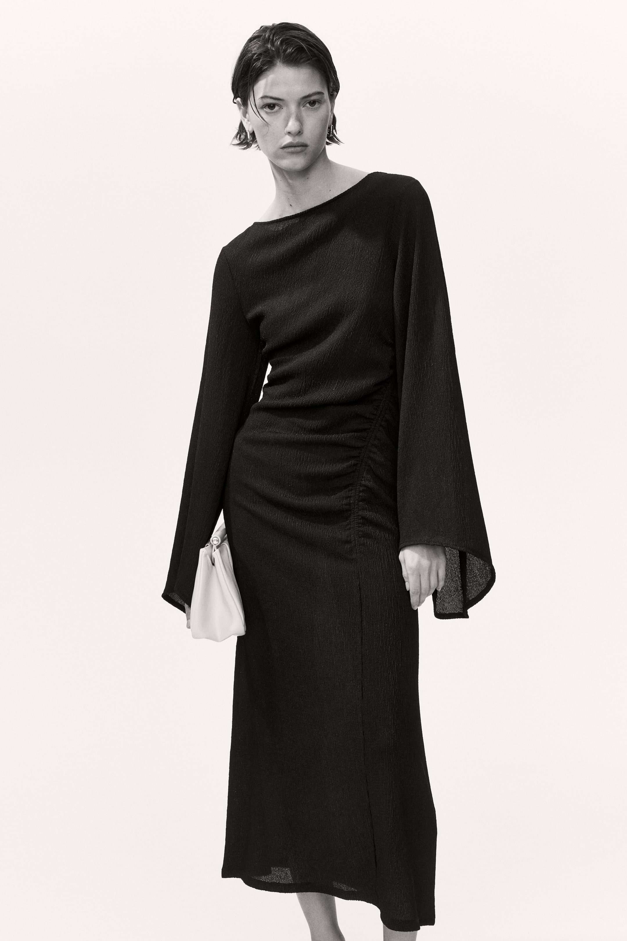 View larger image: This black crinkled dress showcases a wide boat neckline and long, flowing sleeves. The dress cinches at the waist with vertical ruching on the left side, and features a midi-length skirt with a vertical slit on the left.