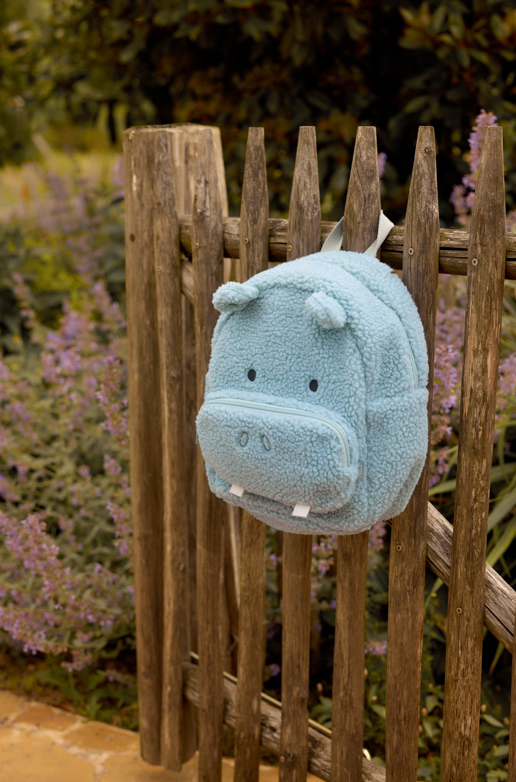 Een lichtblauw-grijze sherpa-fleece kinder-rugzak staat rechtop in een vooraanzicht en is ontworpen om op een nijlpaard te lijken. De rugzak heeft een geweven draaglus aan de bovenkant, kleine afgeronde oren, geborduurde zwarte ogen en een voorvak met ritssluiting, afgewerkt met geborduurde neusgaten en twee witte vilten tanden.