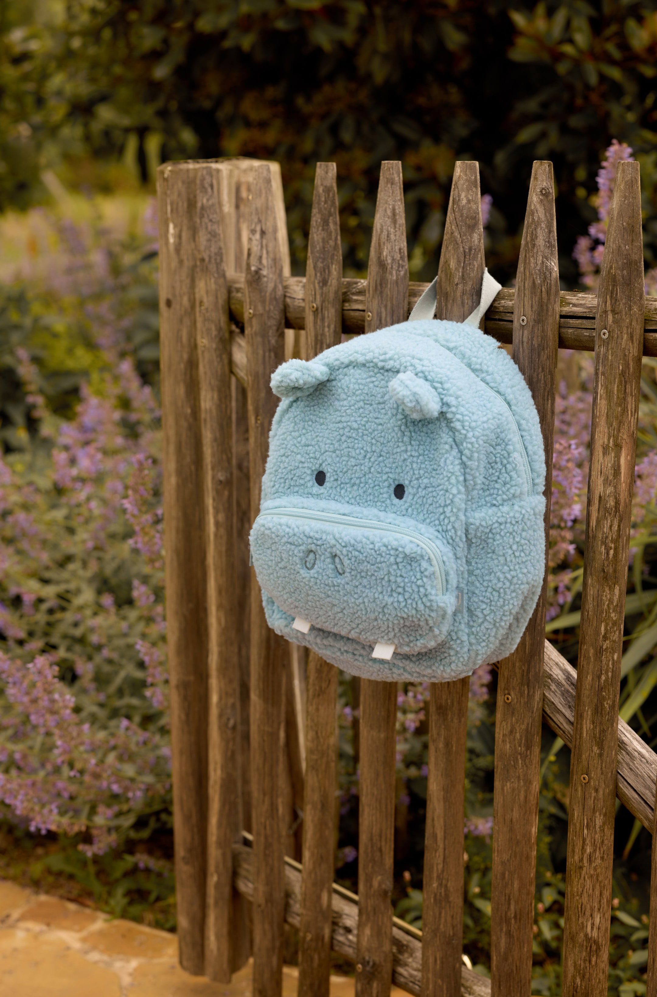 Agrandir l'image: Un sac à dos sherpa bleu clair, conçu en forme d’hippopotame, est suspendu à une clôture en bois, mettant en valeur sa matière polaire texturée sous un angle à trois quarts. Le sac à dos présente de petites oreilles arrondies, des yeux brodés et une poche avant zippée en forme de museau, avec deux dents en tissu blanc dépassant du bas.
