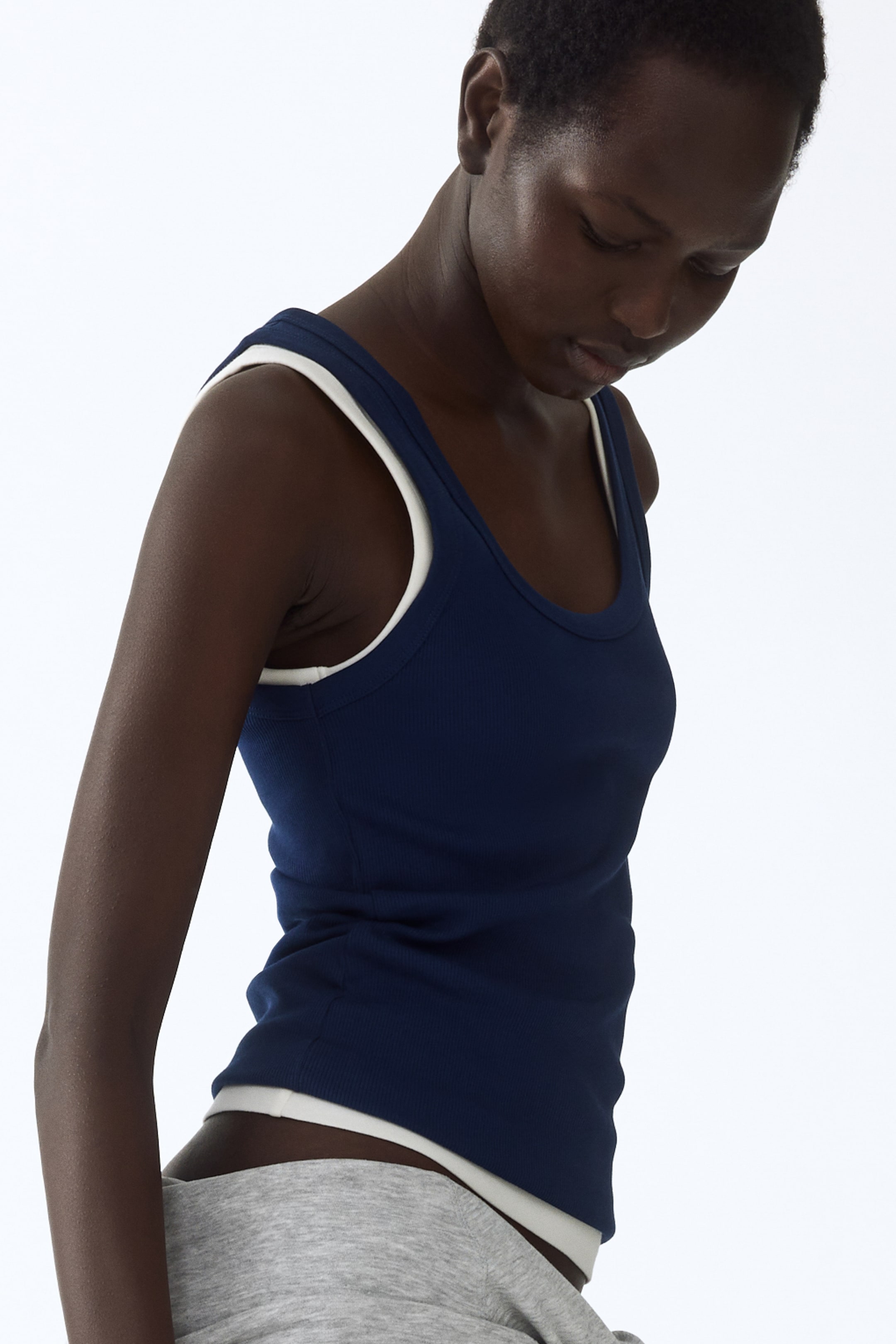 View larger image: The navy blue ribbed knit tank top is shown from an angled side-front perspective, showcasing a fitted silhouette, a scoop neck, and white trim visible along the neckline, armholes, and hem.