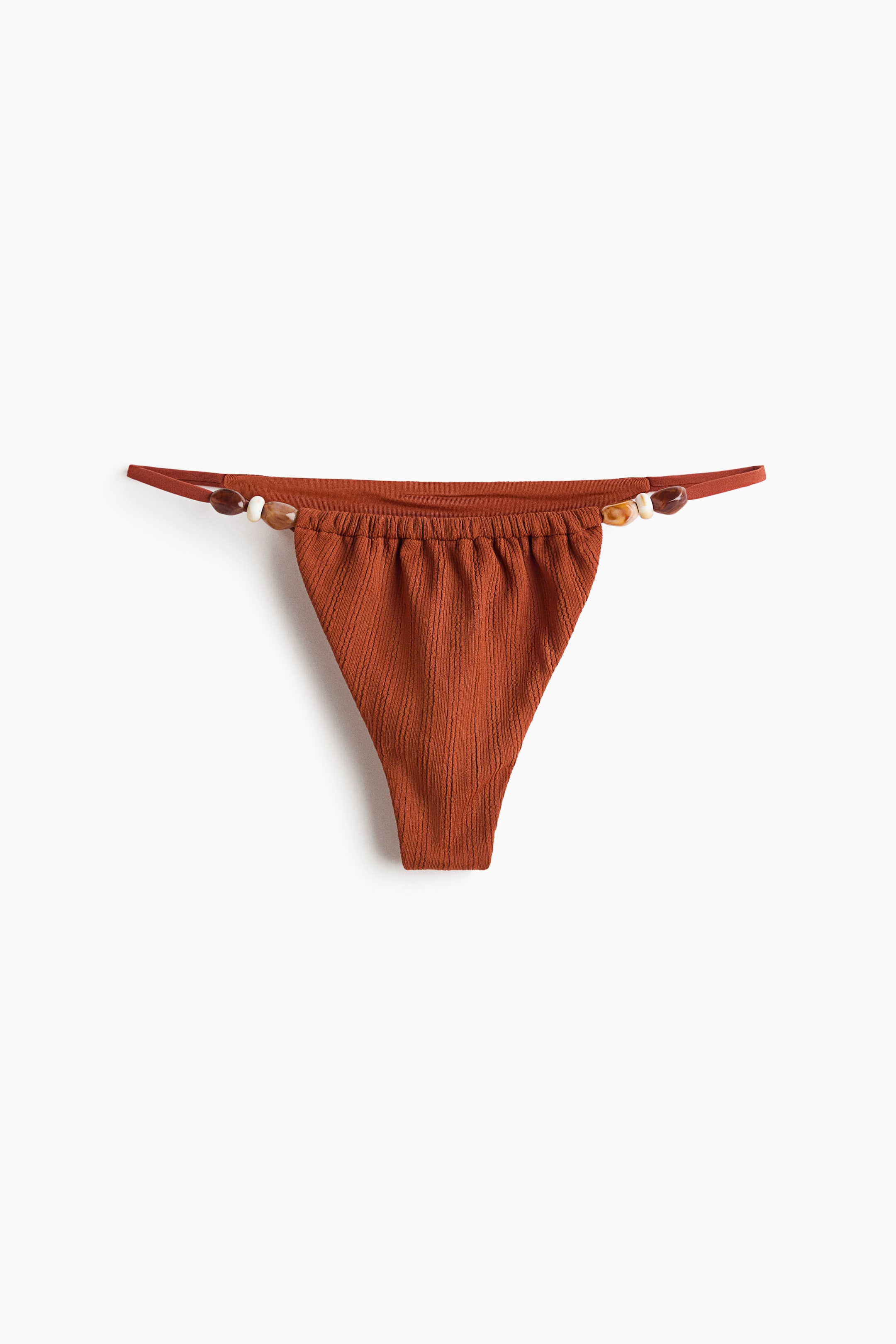 Vezi imaginea mai mare: Un slip de bikini portocaliu ars este prezentat din față, având un stil tanga cu decupaj înalt, realizat dintr-un material cu textură subtilă și cu marginea de sus adunată. Curele laterale subțiri se extind de la partea superioară, fiecare fiind împodobită cu mai multe mărgele decorative în tonuri de alb, maro și chihlimbar.