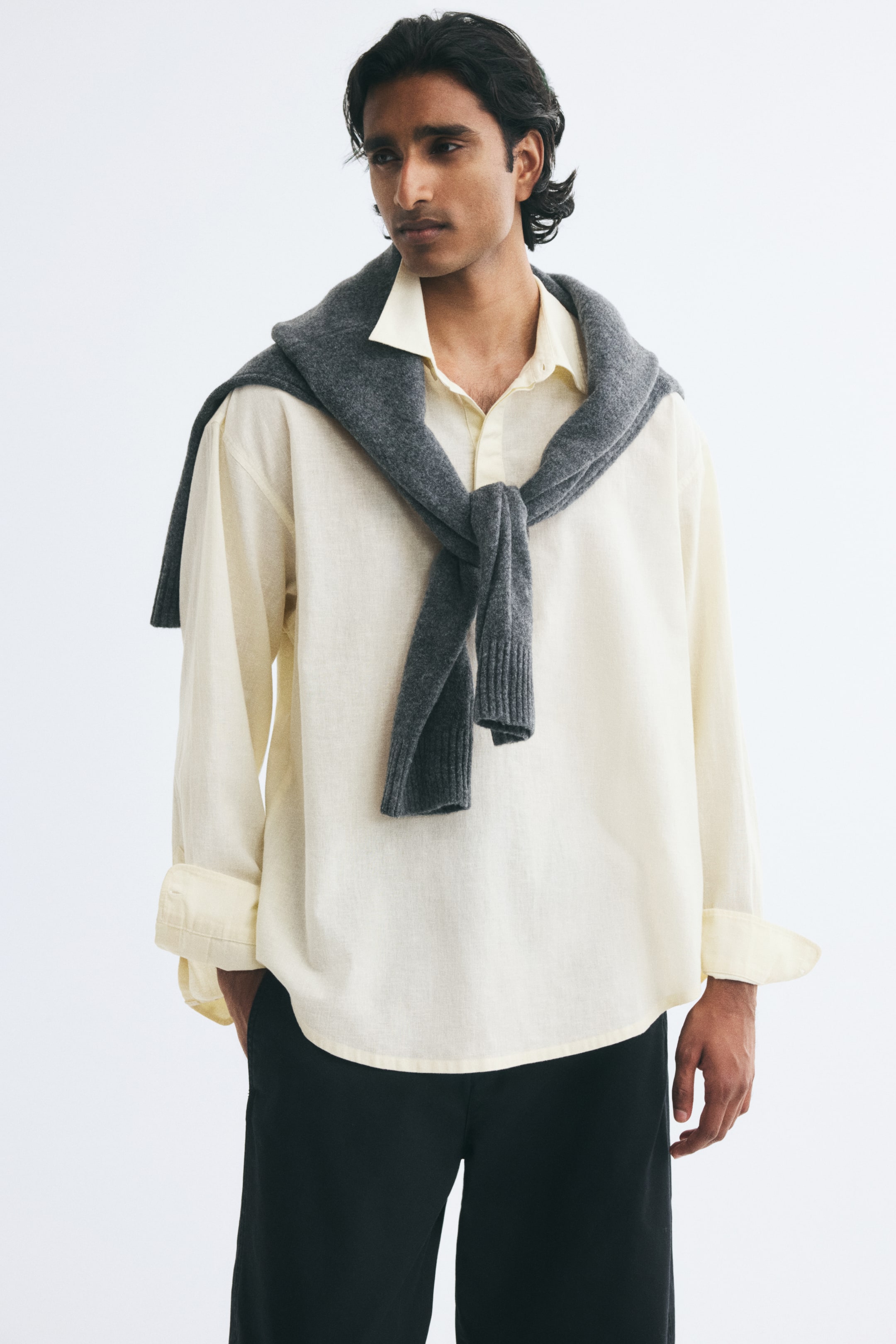 View larger image: A light yellow linen-blend popover shirt is being worn by the model, featuring a relaxed fit, dropped shoulders, an open collar, and rolled-up cuffs. The shirt is styled with a grey knit sweater draped over the shoulders and paired with dark trousers.