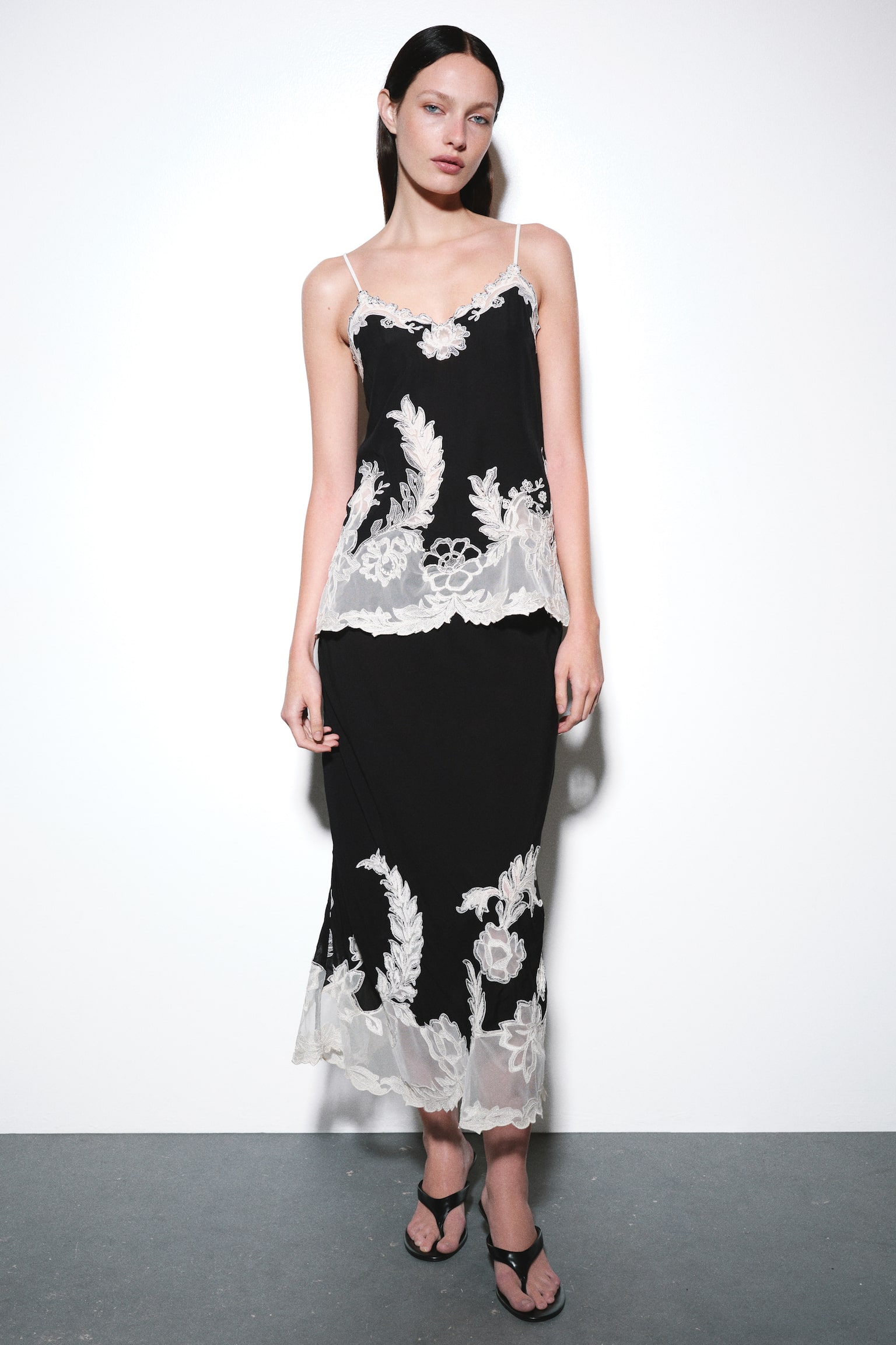 A black midi skirt is shown from the front, featuring a smooth, straight silhouette. The lower portion of the skirt is decorated with intricate cream-colored lace appliquï¿½ in floral and leaf designs, extending upward from a sheer, scalloped hem. - 1