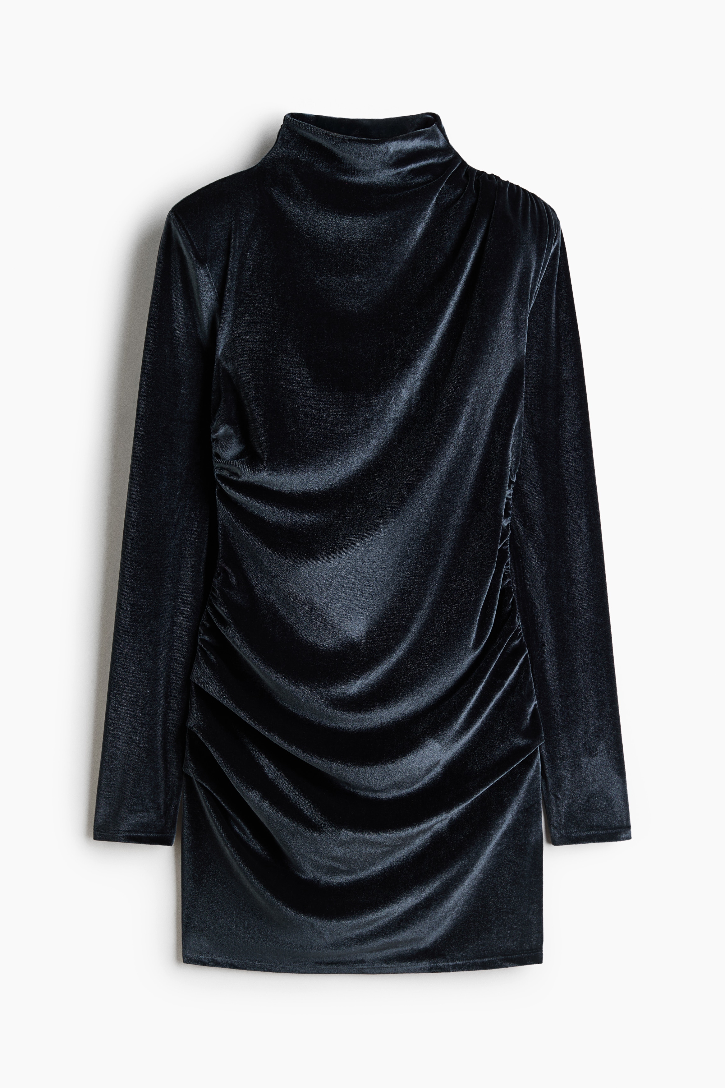 Une mini-robe moulante en velours noir est portée par le mannequin, avec un col montant, des épaules structurées à épaulettes et des manches longues. Le modèle présente des coutures froncées sur l’ensemble, créant un effet drapé, et est stylé avec des collants noirs transparents.