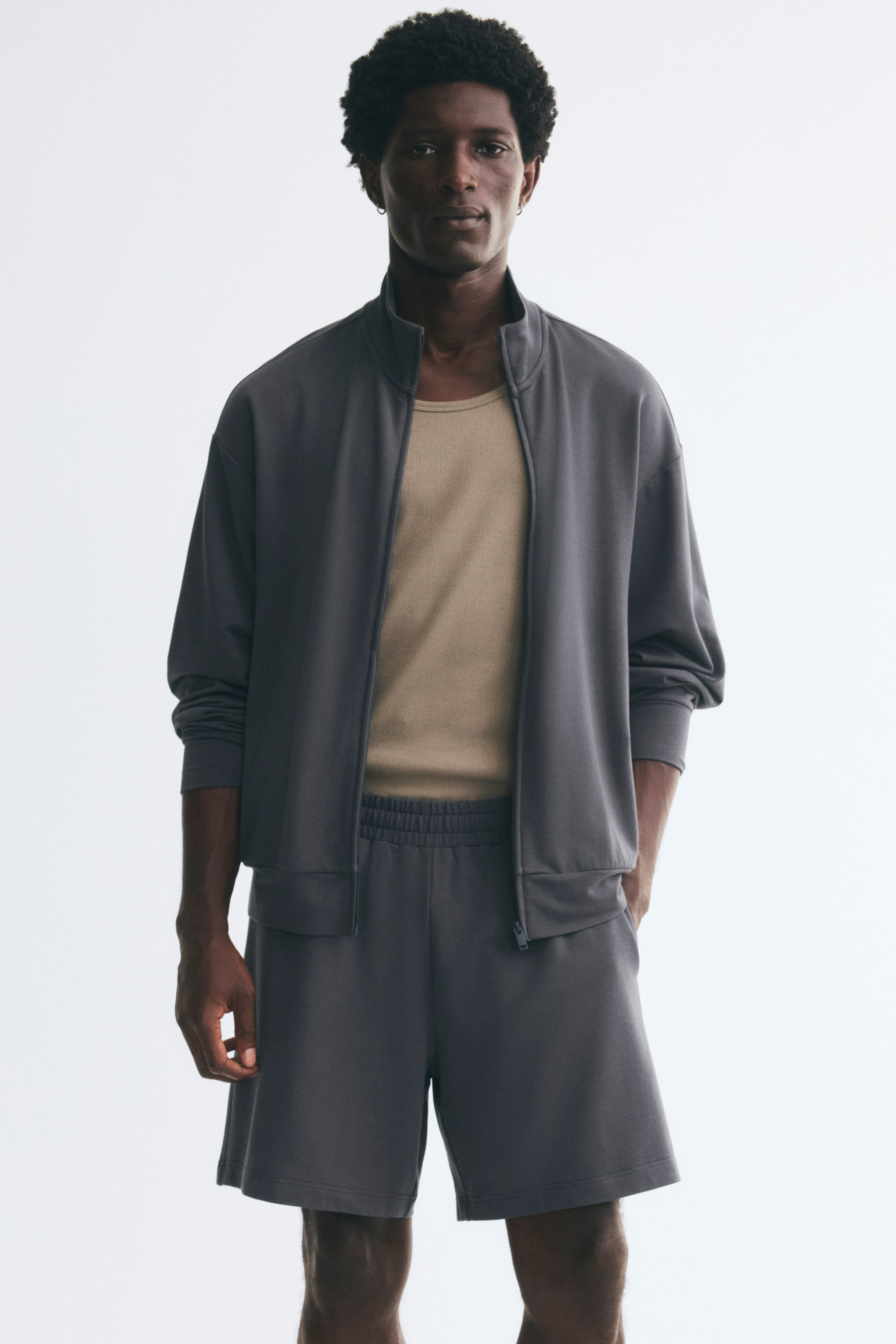 View larger image: Dark gray sweat shorts featuring an elastic waistband and a relaxed fit are worn by the model. The shorts are styled with a matching zip-up jacket and a tucked-in beige top, with the model resting one hand in a side pocket.