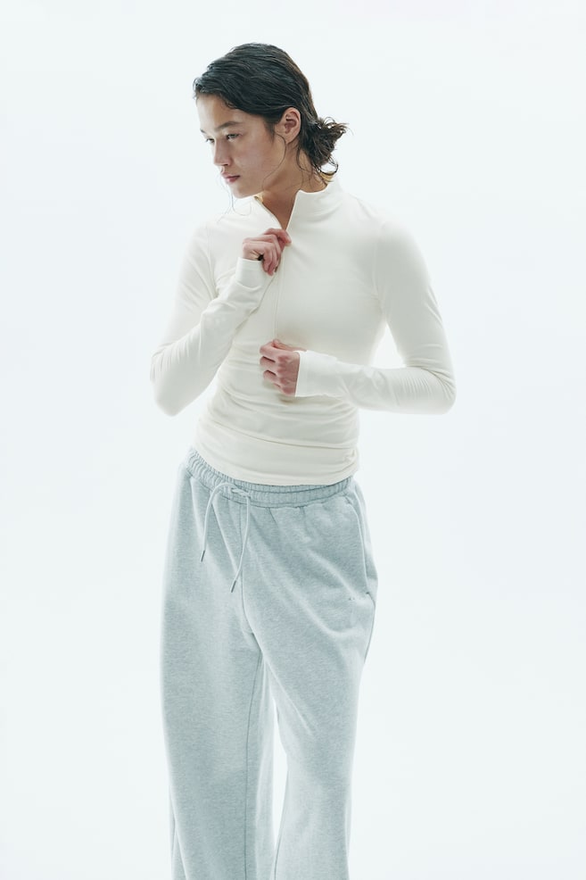 La modella indossa un top color bianco sporco, a maniche lunghe, con mezza zip, mentre regola la zip sul colletto. Il top aderente è abbinato a pantaloni della tuta grigio chiaro con coulisse. - 1