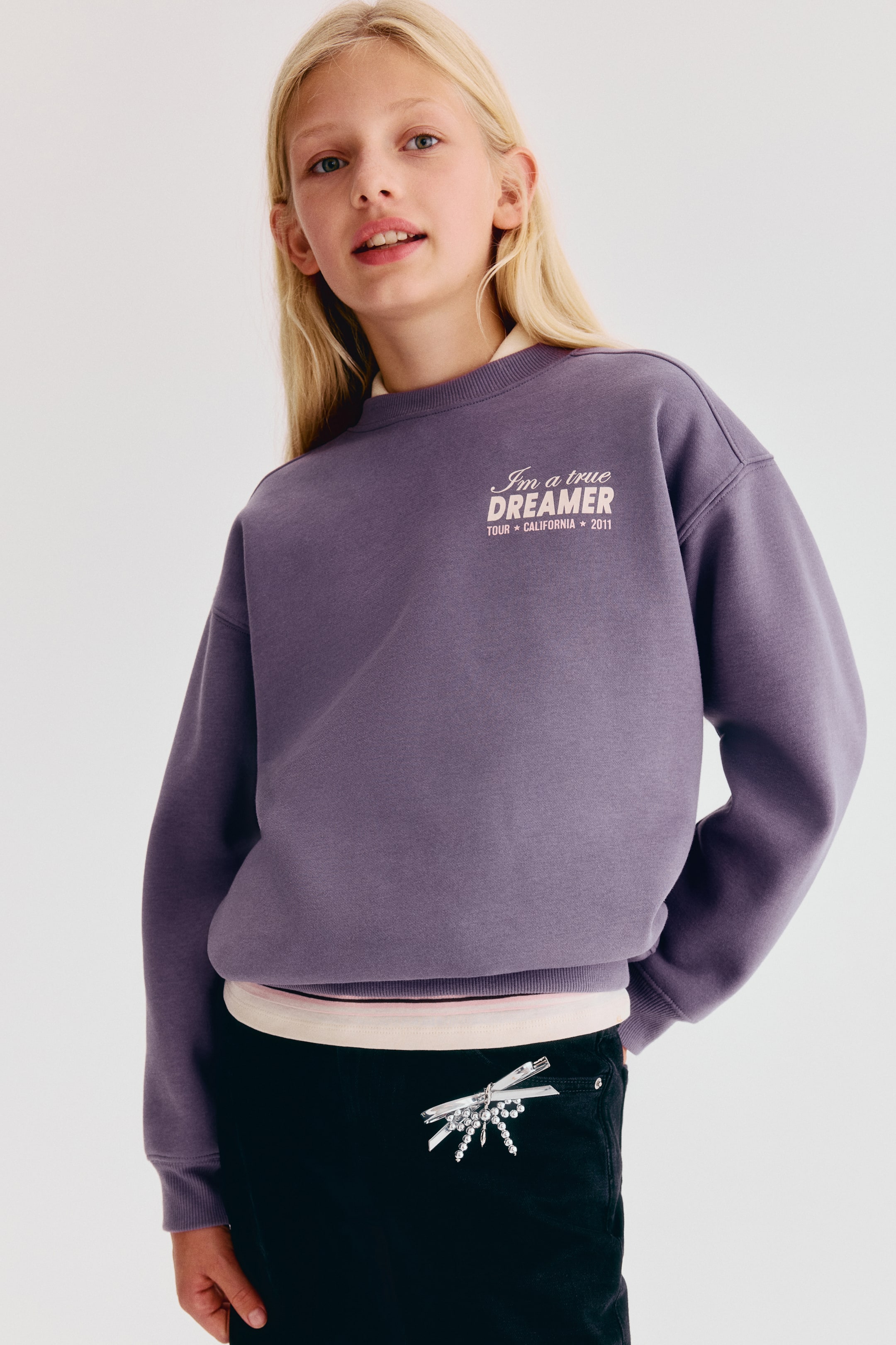 Agrandir l'image: Un sweat-shirt ample violet en tissu polaire gratté est présenté de face, avec un col rond bordé de bord-côtes, des épaules tombantes et des manches longues à poignets côtelés. Le sweat-shirt affiche un motif texte imprimé sur la poitrine gauche indiquant « I'm a true DREAMER » et est porté par-dessus un haut de couleur claire visible au niveau de l’encolure et de l’ourlet.