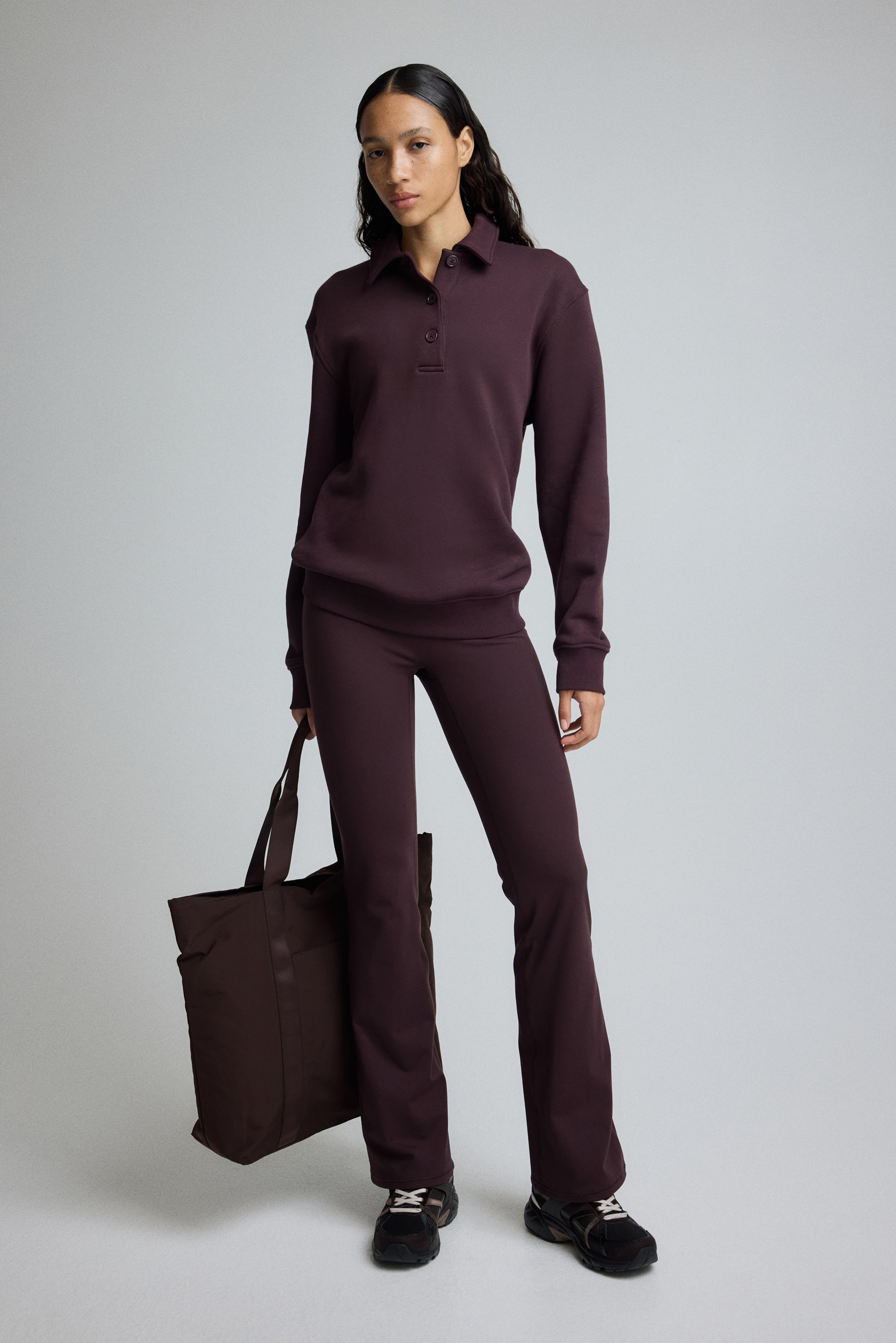 Grotere afbeelding bekijken: Een bijpassende set in donker pruim wordt frontaal gepresenteerd, bestaande uit een polotrui en uitlopende broek. De trui heeft een kraag, een knoopsluiting met drie knopen en geribde boorden aan de mouwen en zoom, en de broek sluit aan op de dijen en loopt iets uit naar de enkel. Een bijpassende donker pruimen draagtas wordt aan de zijkant vastgehouden.