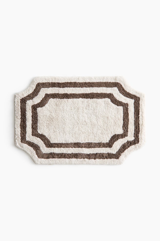An ornate bath mat presents a creamy white plush texture. It features a scalloped edge defined by a dark brown border, with a second parallel dark brown stripe outlining the creamy white central area. - 1