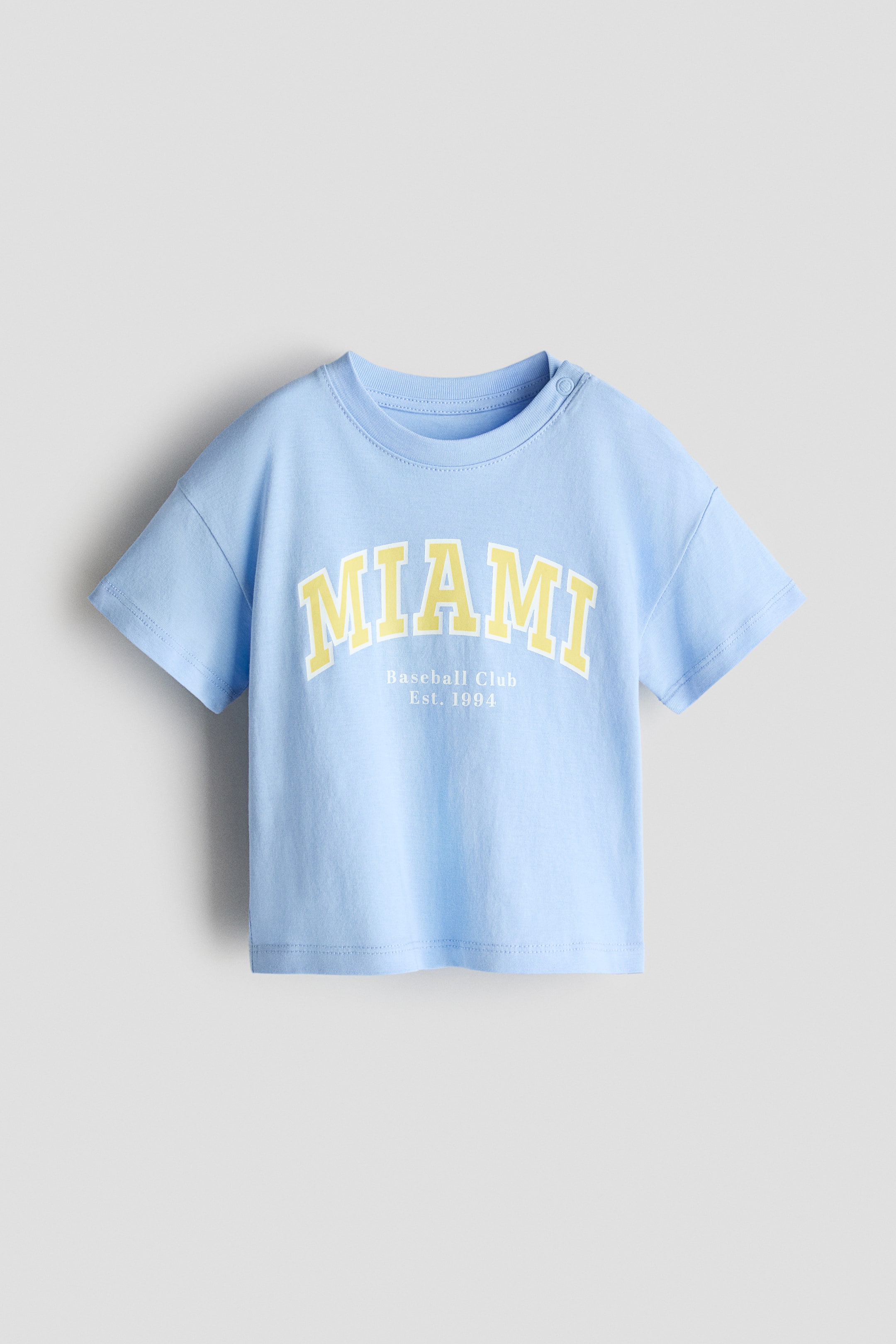הצגת תמונה גדולה יותר: חולצת טי מכותנה בצבע כחול בהיר מוצגת במבט קדמי, עם צווארון עגול מצולע ושני כפתורי לחיצה על הכתף השמאלית. בחזית מופיע הכיתוב "MIAMI" באותיות קשתיות בצבע צהוב עם קו מתאר לבן, יחד עם הכיתוב "Baseball Club Est. 1994" באותיות קטנות בצבע לבן מתחת.