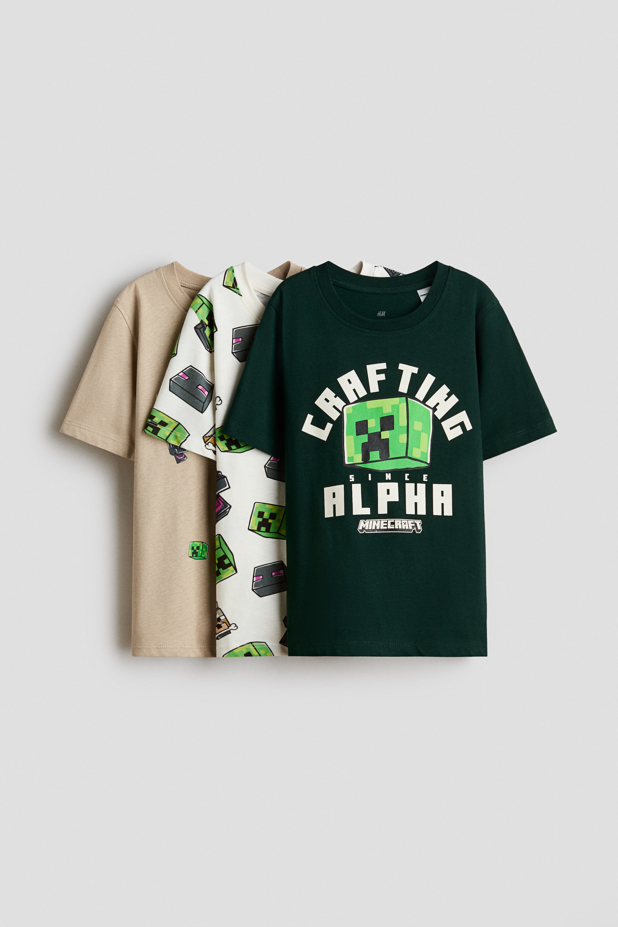 View larger image: Three classic cotton jersey T-shirts are shown facing forward in a layered arrangement, featuring ribbed necklines. The front dark green T-shirt displays a printed Minecraft graphic, the middle off-white T-shirt features an all-over character print, and the back T-shirt is shown in solid light beige.