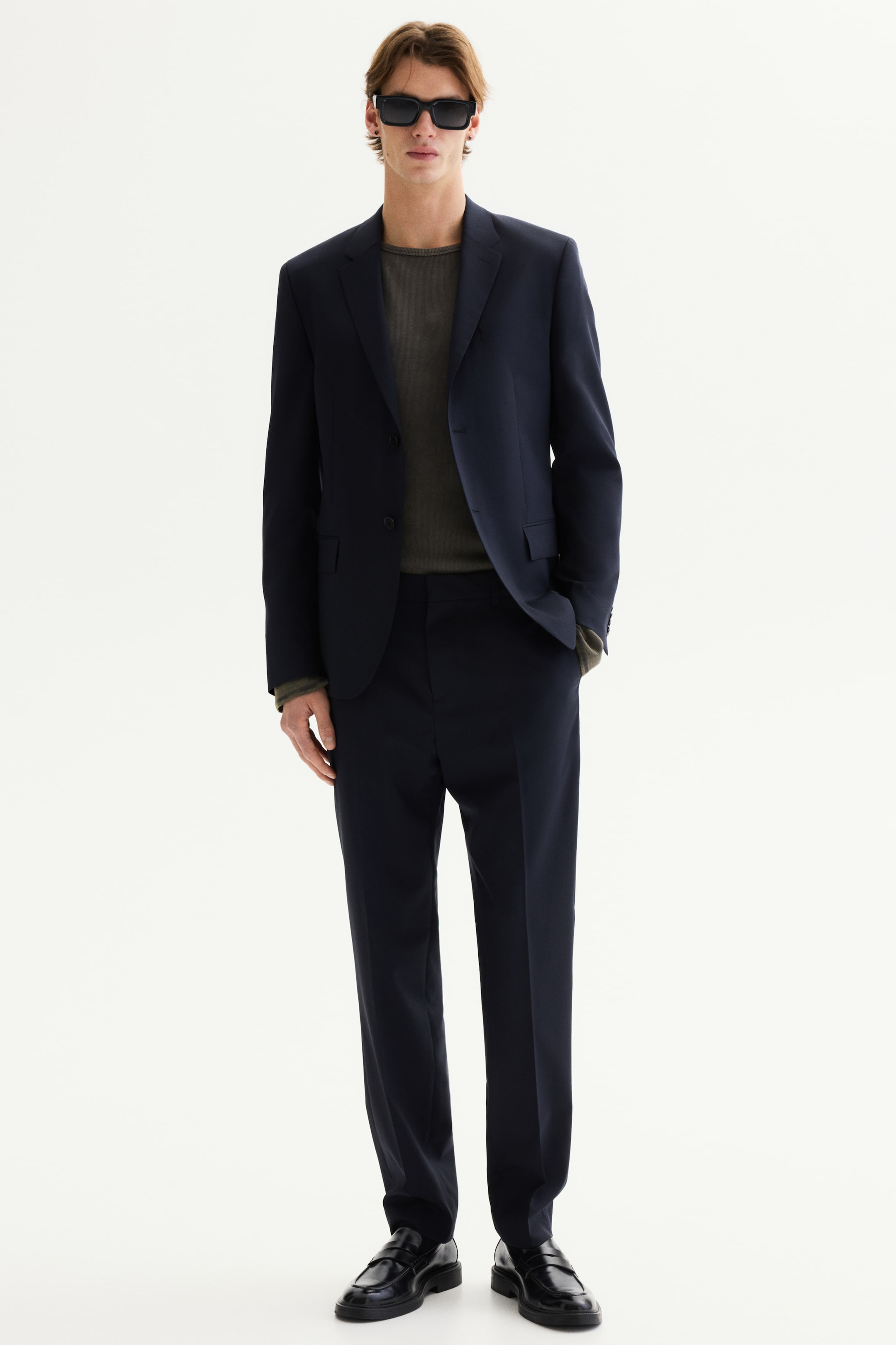 View larger image: A navy blue suit is showcased, featuring a single-breasted jacket with notch lapels, a two-button closure, and flap pockets, paired with matching straight-fit trousers. The jacket is styled over a dark olive long-sleeved top, highlighting its fit and structure.