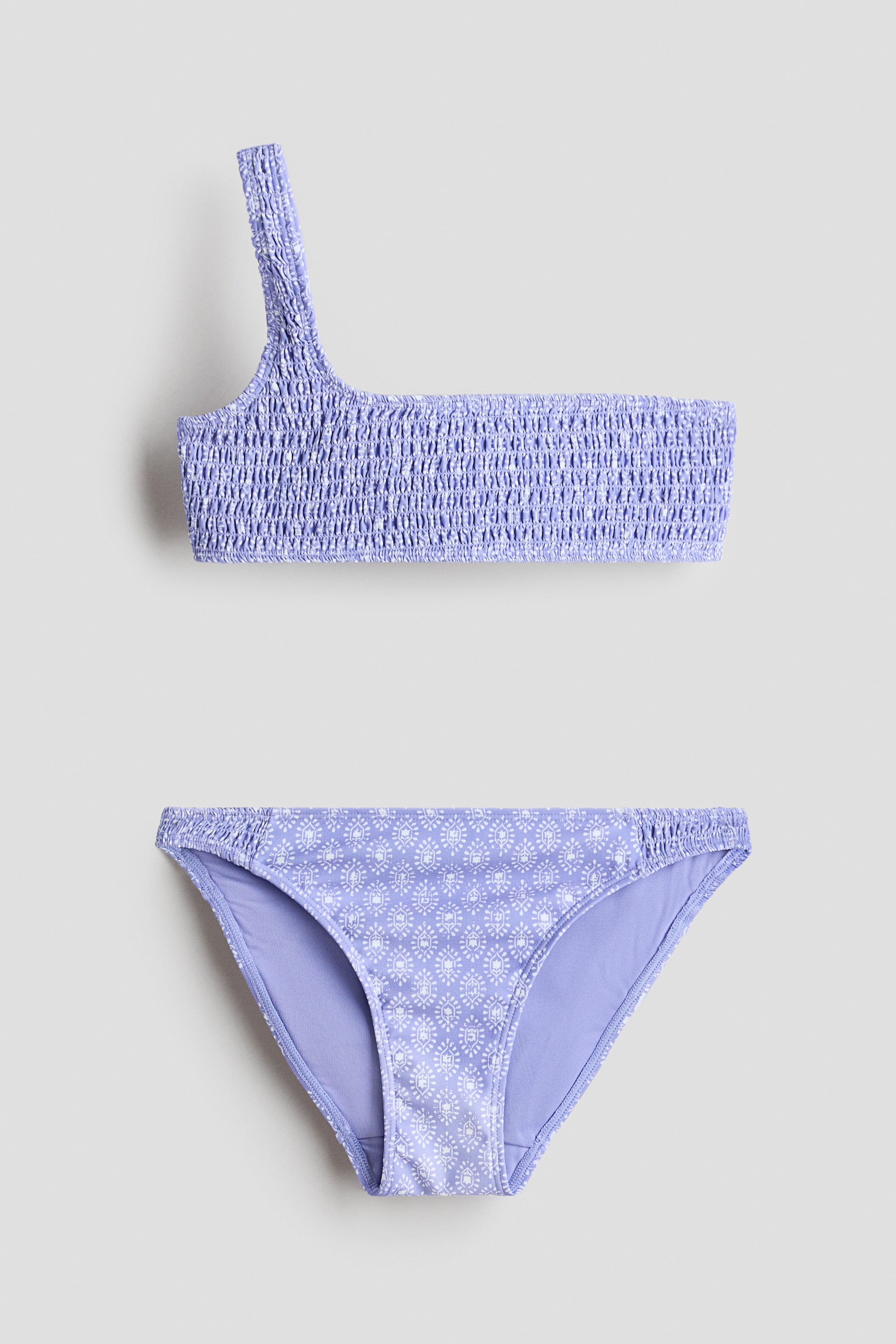 Grösseres Bild ansehen: Zweiteiliges Badeanzug-Set mit einem schulterfreien, gesmokten Bandeau-Top und einem gemusterten Bikiniunterteil. Das lavendelfarbene Top ist komplett gesmokt, und das dazu passende Unterteil präsentiert ein weißes geometrisches Motiv auf lavendelfarbenem Grund.