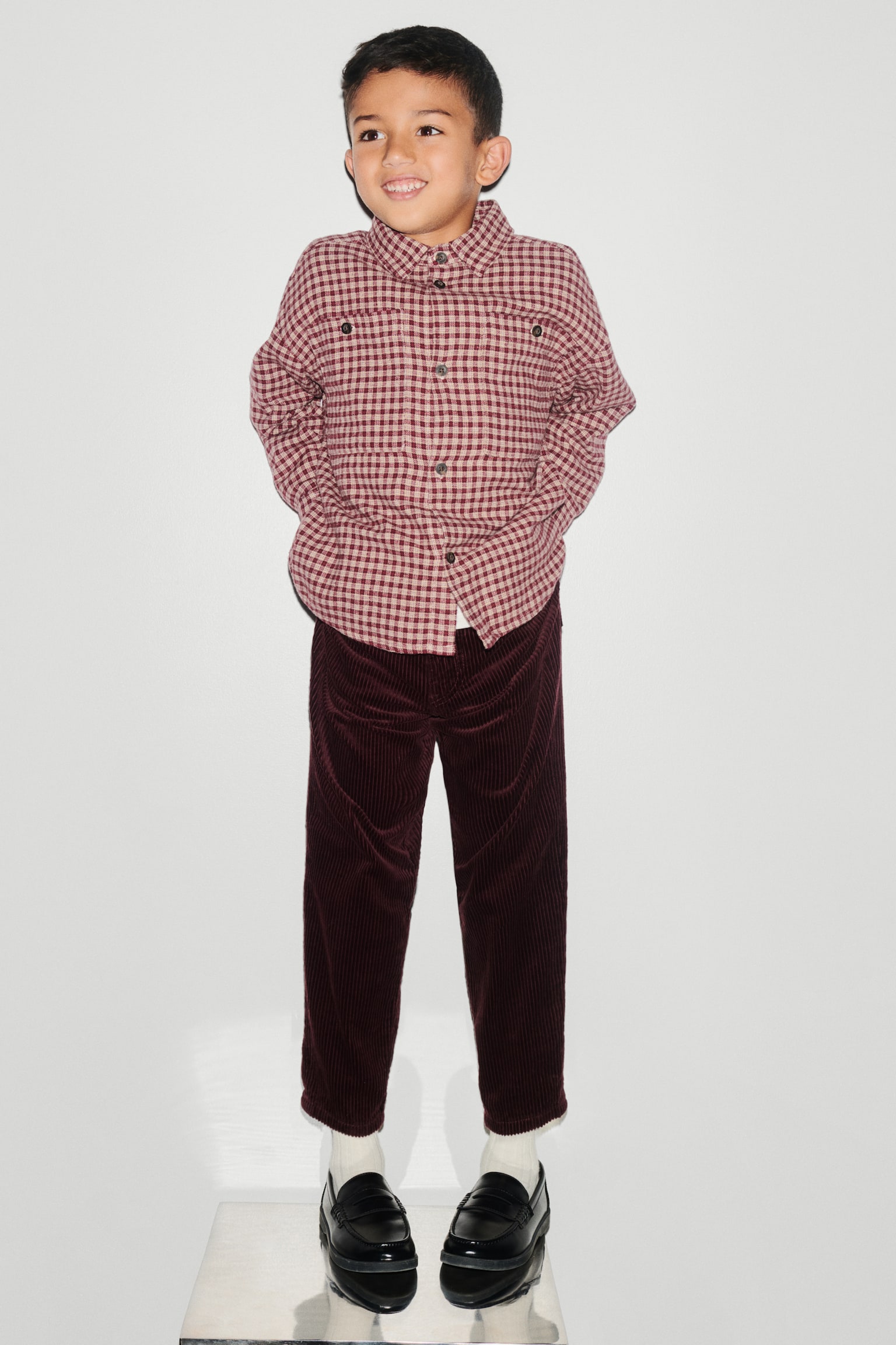 A long-sleeved shirt jacket is shown from the front, featuring a red and cream check pattern. The jacket has a classic point collar, a button-down front with dark contrast buttons, dropped shoulders, and two chest pockets with button closures. - 1