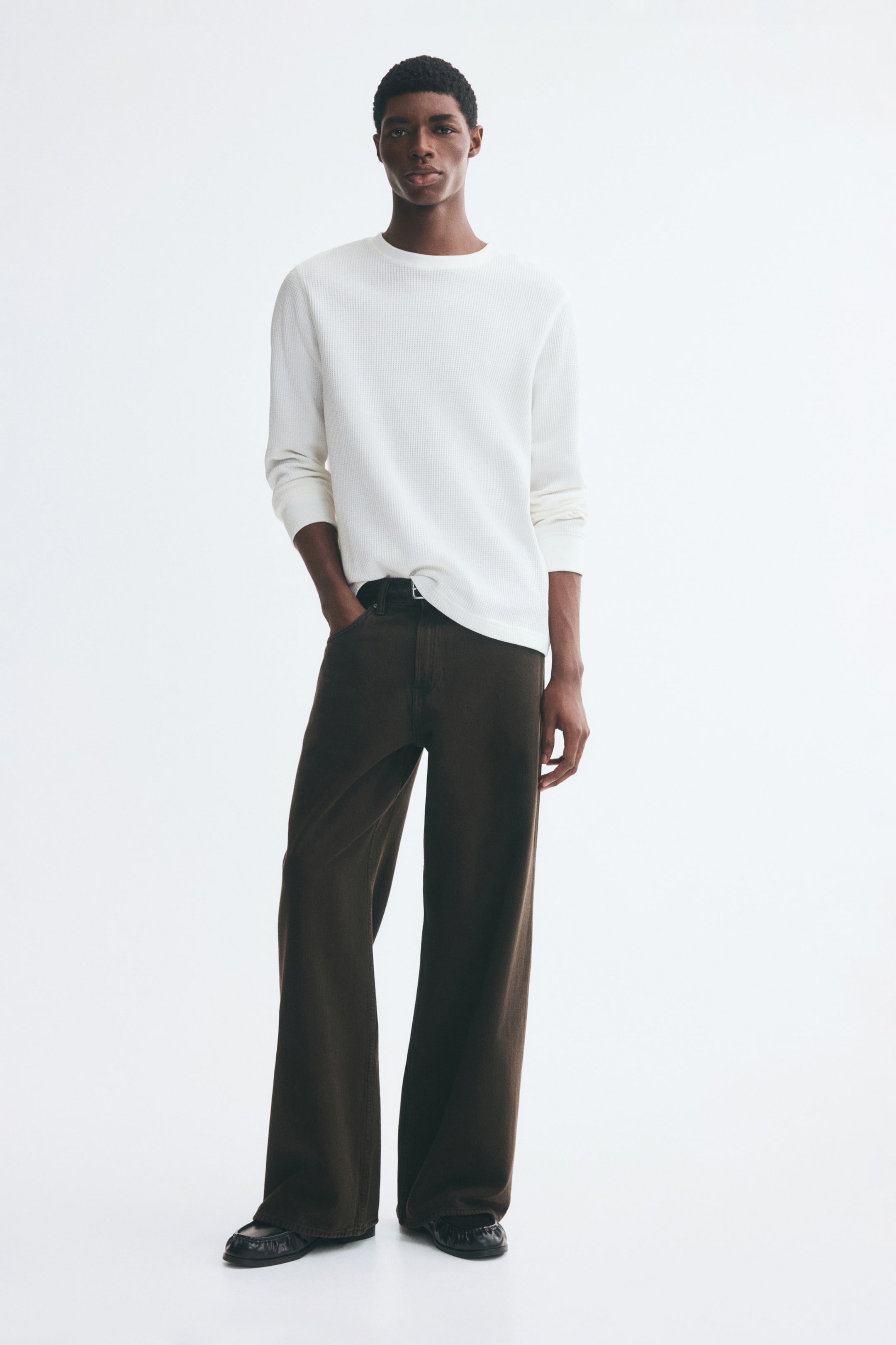 Voir une image plus grande: Un t-shirt blanc à manches longues en jersey de mélange coton texturé gaufré est porté par le mannequin, qui se tient avec une main dans la poche d’un pantalon ample brun foncé à jambes larges. Le t-shirt présente une coupe décontractée, un col rond côtelé et des poignets côtelés.