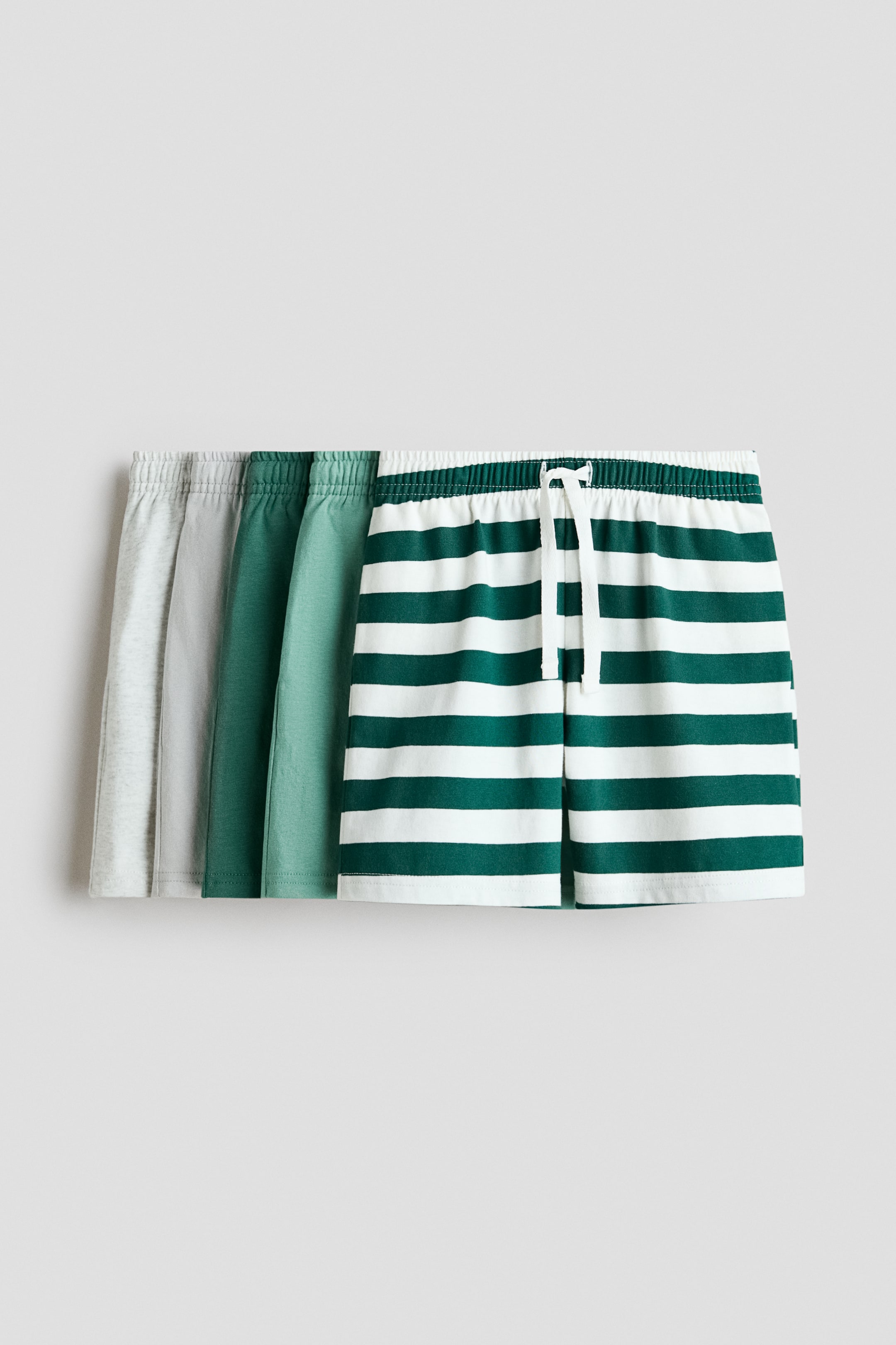 Grotere afbeelding bekijken: Drie paar zachte katoenen jersey shorts worden aan de voorkant gepresenteerd, elk met een elastische tailleband met trekkoord. De shorts overlappen van links naar rechts, waarbij het meest linkse paar lichtgrijs is, het middelste paar effen donkergroen, en het meest rechtse paar witte en donkergroene horizontale strepen vertoont met een wit trekkoord dat aan de voorkant is vastgebonden.