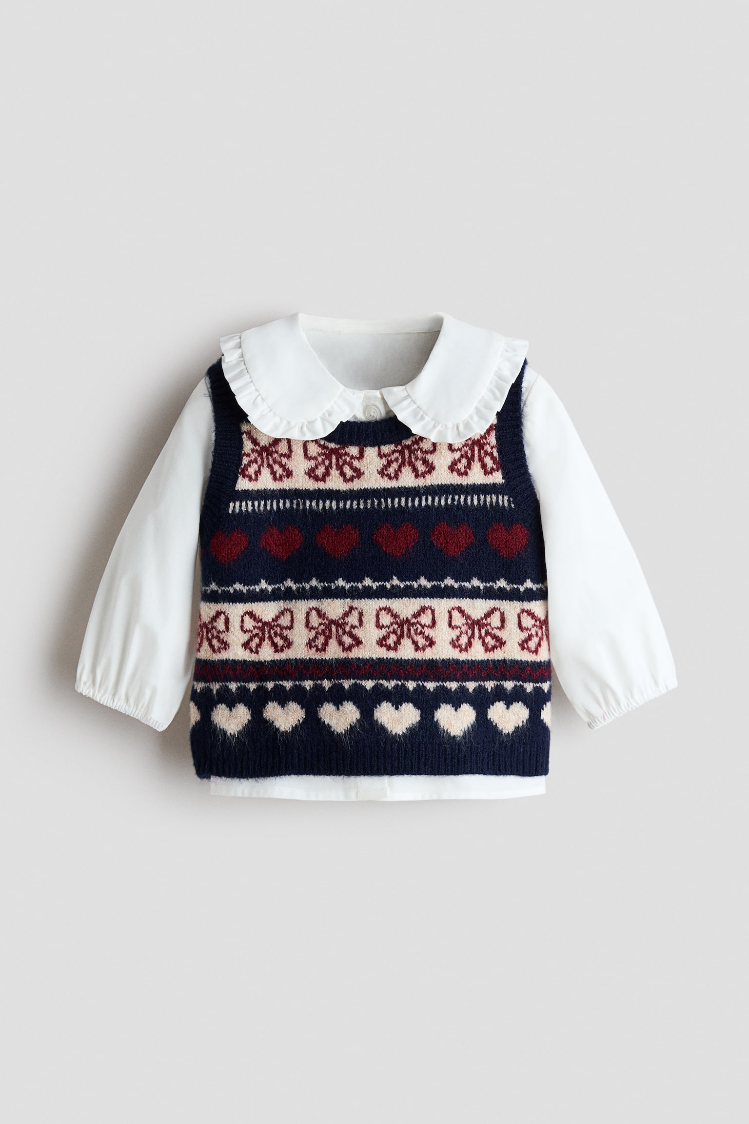 A two-piece set is shown from the front, featuring a white cotton blouse layered beneath a navy blue jacquard-knit sweater vest. The blouse displays a rounded, frill-trimmed collar and long sleeves with elasticated cuffs, while the vest showcases horizontal bands of red and cream hearts and bows with ribbed trims at the neckline, armholes, and hem.