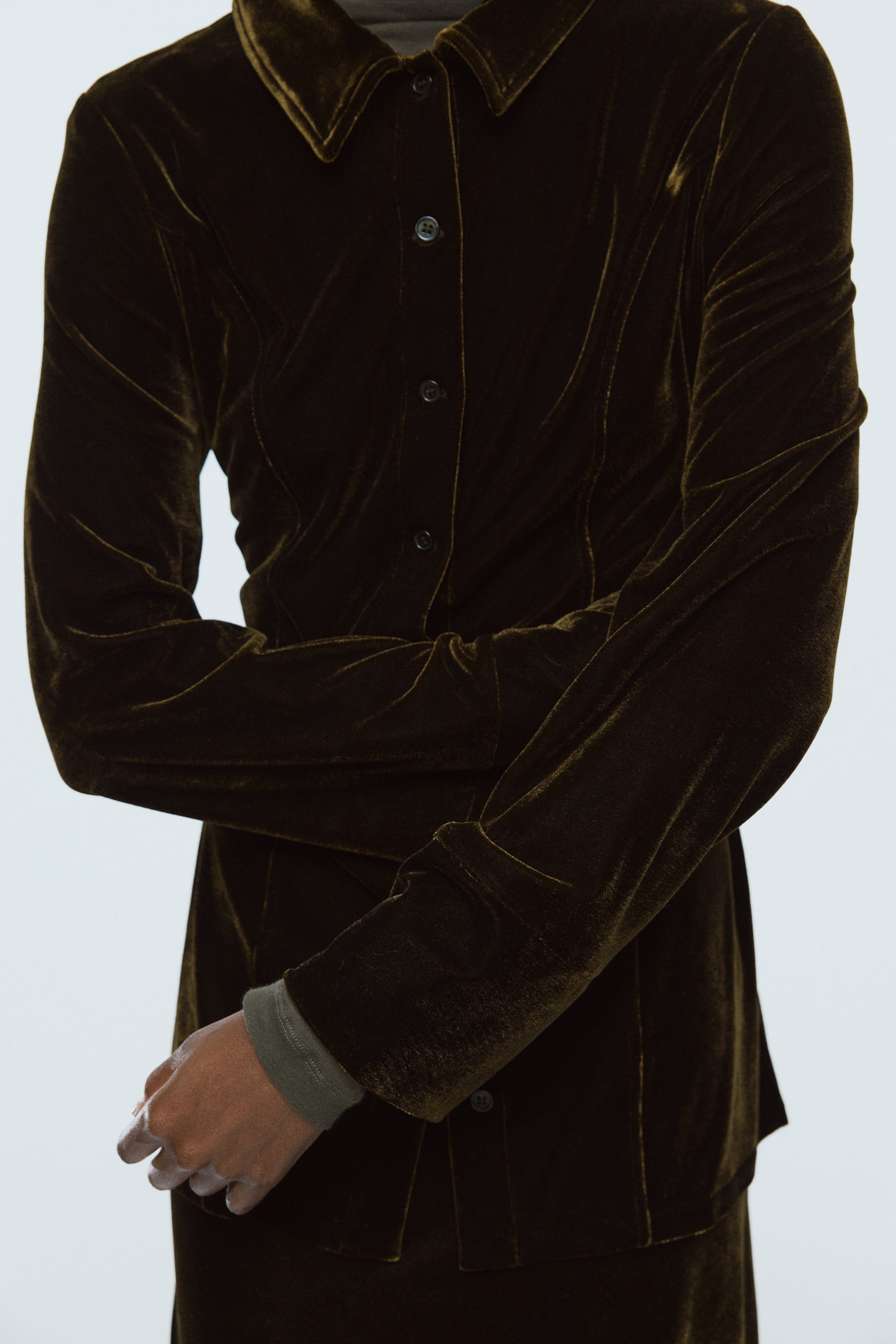 View larger image: A dark olive green velvet button-up shirt is being presented from the front with arms crossed, featuring a pointed collar and dark buttons down the placket. A lighter olive green ribbed long-sleeved top is showing at the cuffs, layering underneath the velvet shirt and highlighting its long sleeves and tailored fit.