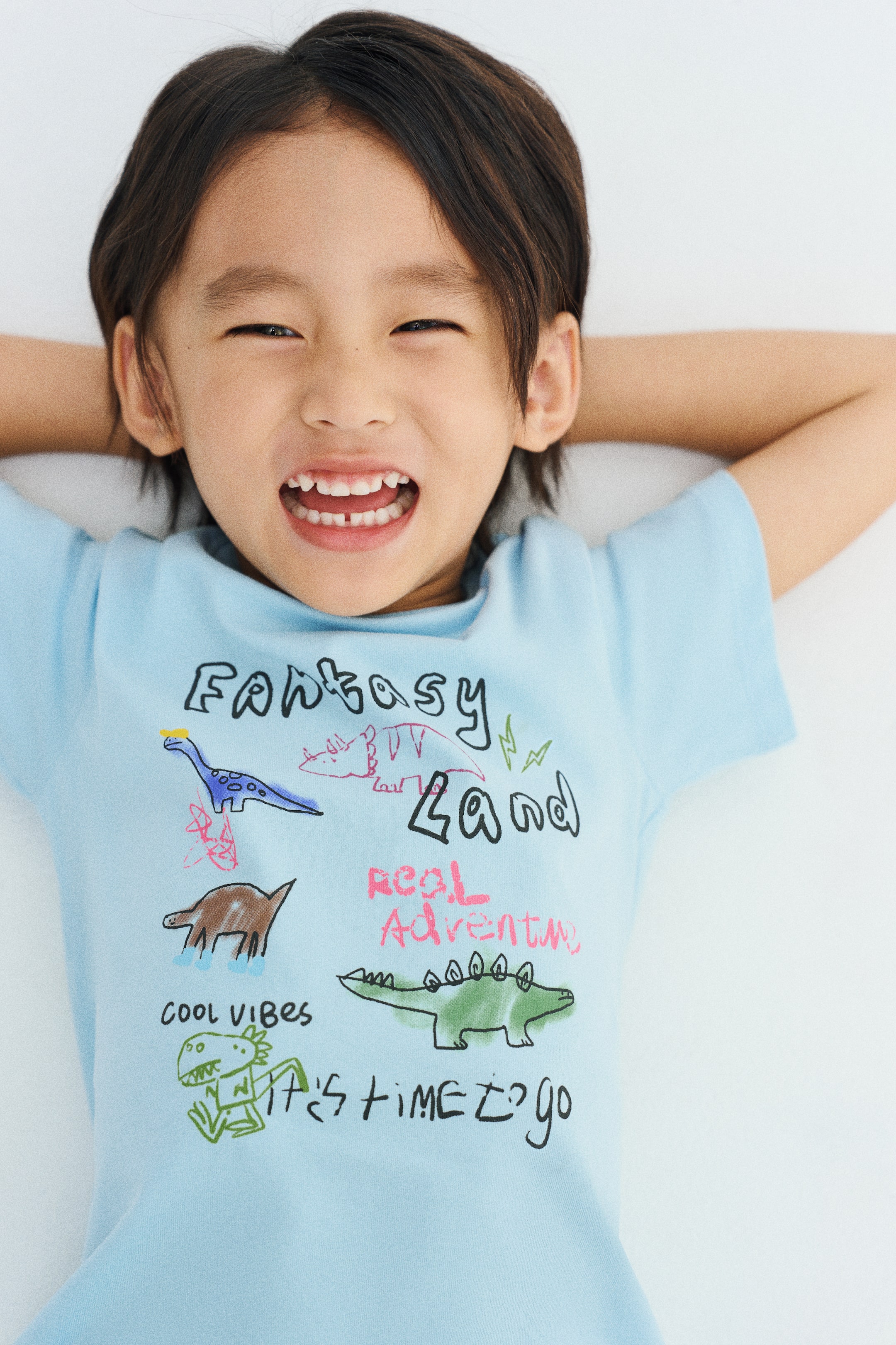 View larger image: A light blue cotton jersey tee featuring a round, rib-trimmed neckline and a colorful dinosaur printed design is being worn by the model. The model is lying down with arms behind their head, showcasing the front of the shirt.