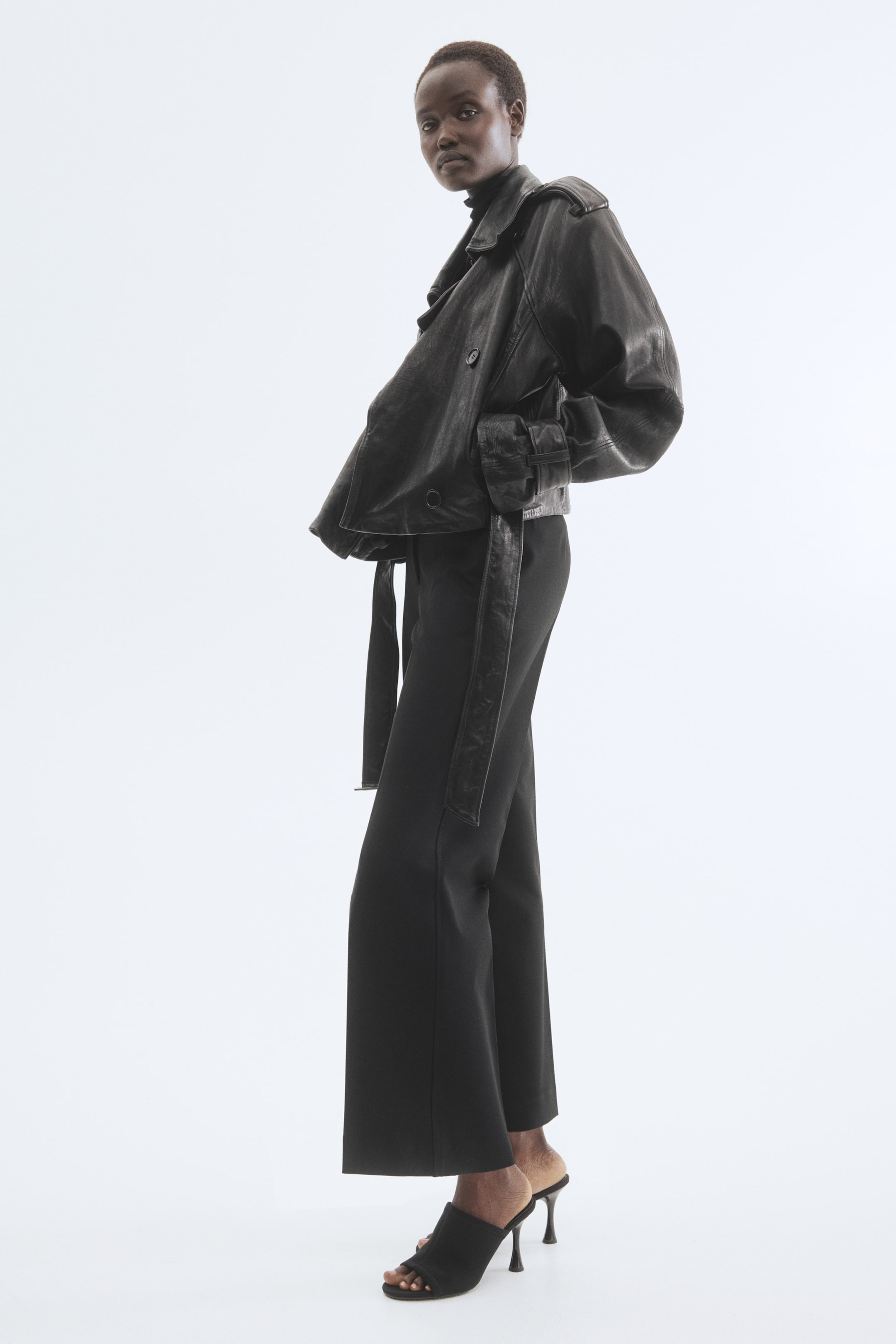 Agrandir l'image: Une veste en cuir noire à double boutonnage présente de larges revers, des épaulettes, et des poignets ceinturés sur des manches volumineuses, avec une ceinture à la taille nouée lâchement. Elle est associée à un pantalon noir à jambes larges et des mules à talons aiguilles ouvertes, le tout présenté sous un angle de trois-quarts.