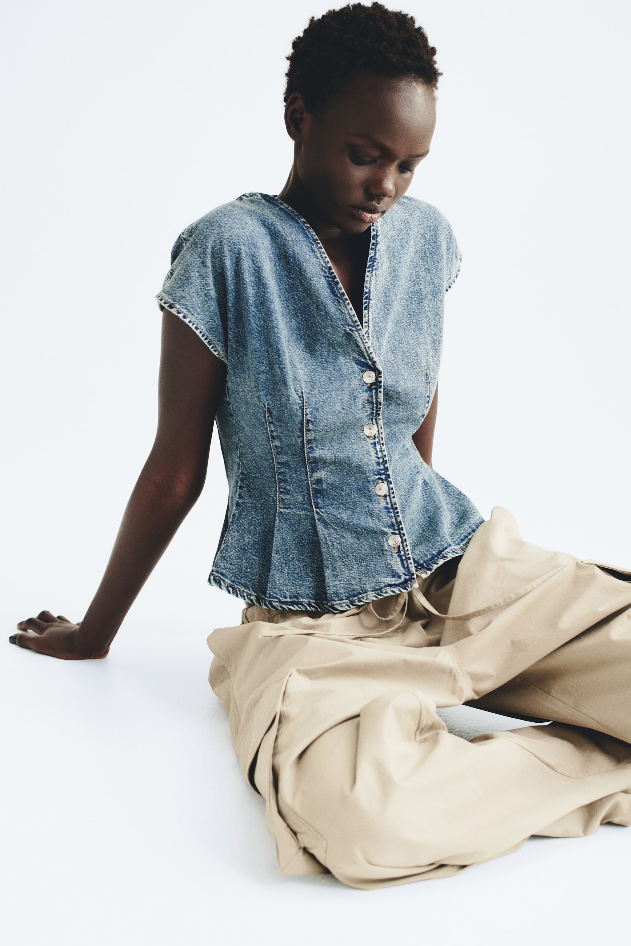 View larger image: The model is wearing a blue cotton denim top featuring a V-shaped neckline, button-down front, cap sleeves, and a flared peplum hem. The top is styled with wide-leg beige cargo trousers.