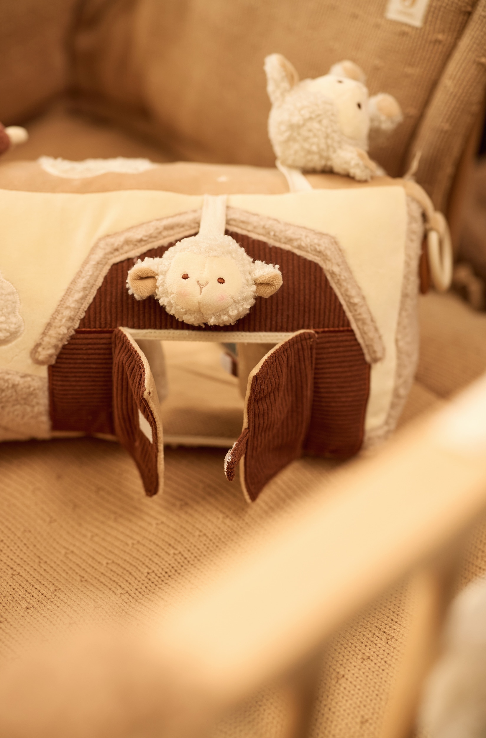 Een cilindervormig baby-activiteitenspeelgoed ligt in een driekwart hoek en laat een mix zien van glad beige textiel en zachte crèmekleurige sherpa-fleece met structuur. Het speelgoed is voorzien van diverse zintuiglijke elementen, waaronder een knuffelzacht schaapje, een bruine halve maan en een plastic bijtring, naast een geborduurd schaapje in het midden en een corduroy symbool.