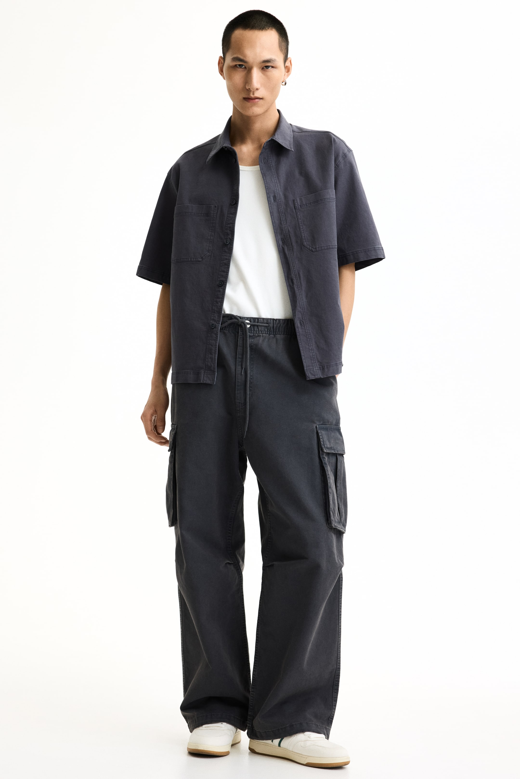 View larger image: A charcoal short-sleeved shirt, featuring two chest patch pockets, is worn open over a white ribbed top. It is styled with charcoal wide-leg cargo pants, designed with an elasticated drawstring waist and large side pockets, while white sneakers complete the ensemble.