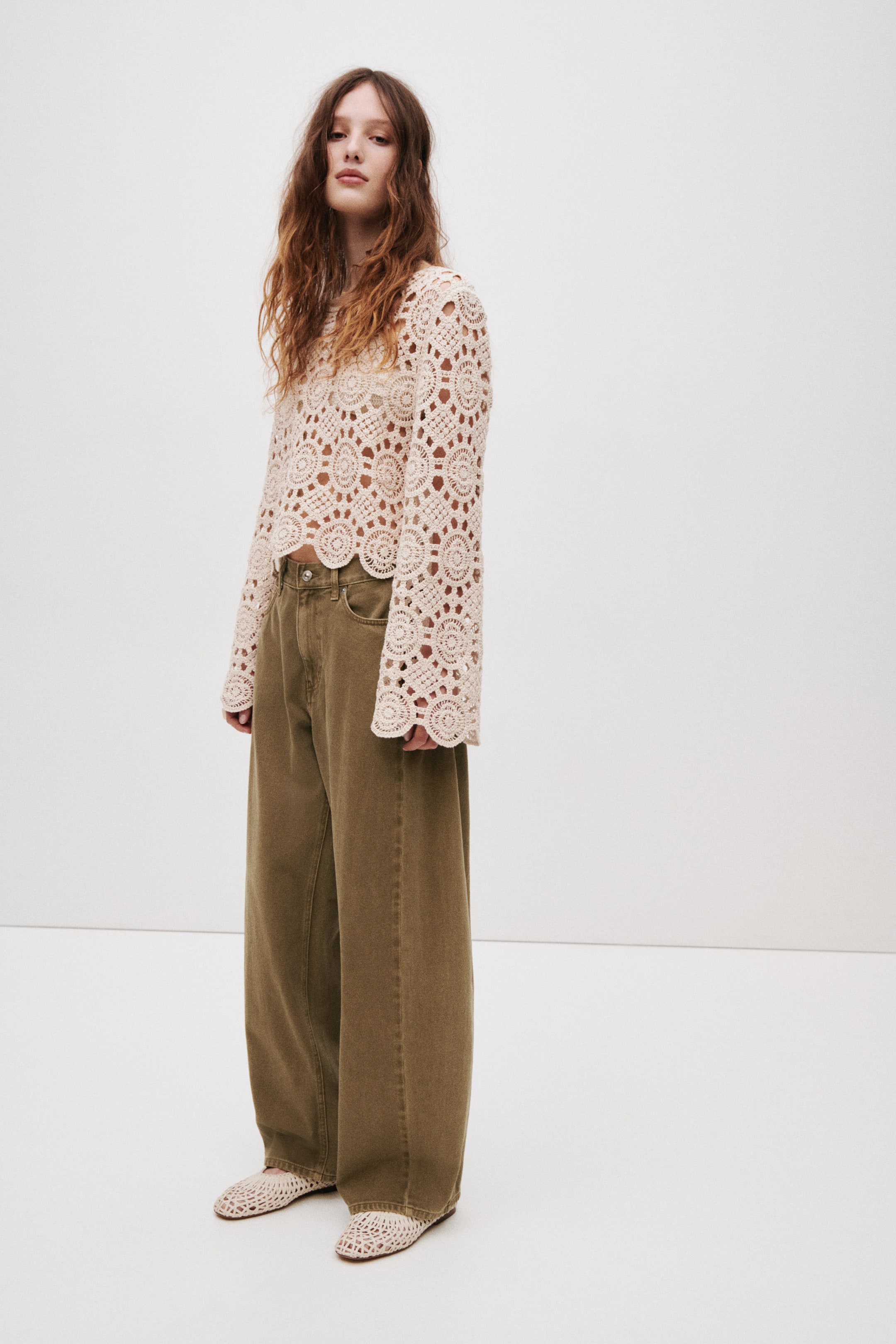 View larger image: The beige crochet top features a round neck and long, flared sleeves, showcasing an intricate open-knit pattern and a scalloped hemline.