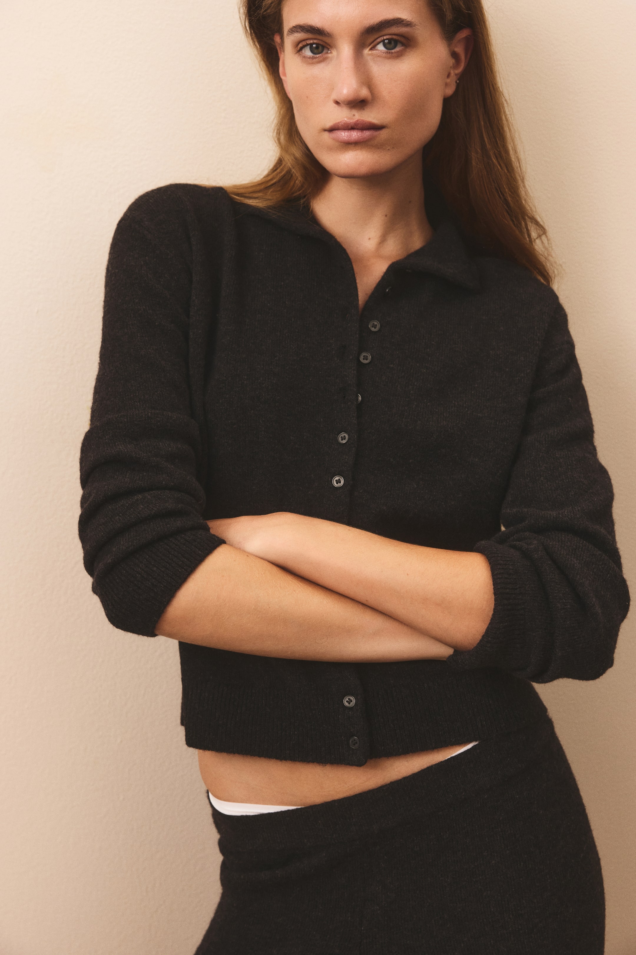 View larger image: A dark grey cardigan in a soft, fine knit is shown from the front, featuring a polo-style collar, a button-down front, and dropped shoulders. The long-sleeved garment has ribbed cuffs and a cropped, ribbed hem, and is styled with matching knit bottoms.