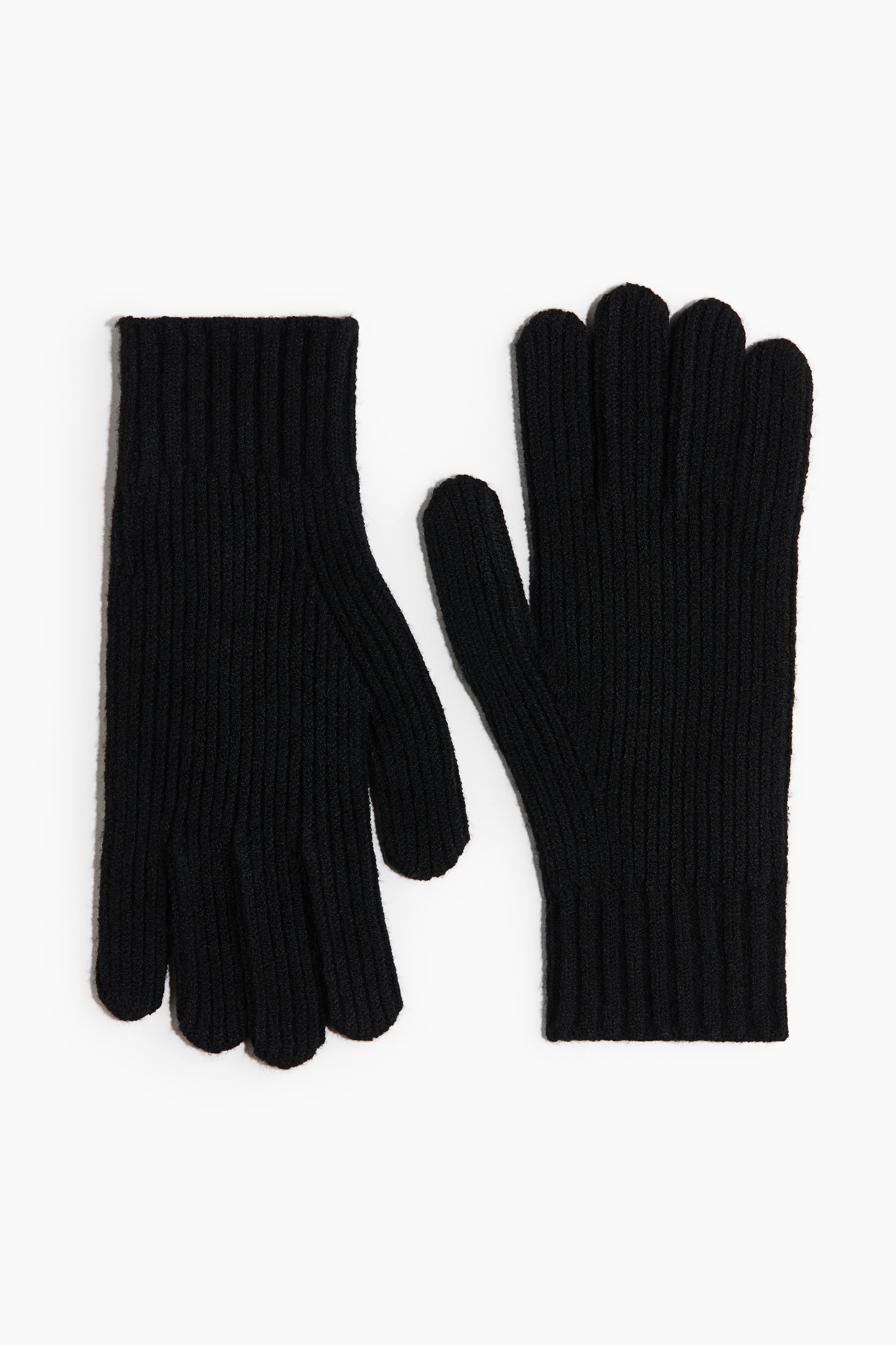 Black gloves in a soft, fine knit with textured ribbed cuffs are being worn by the model, who is posing with arms crossed over a structured black coat. The gloves extend up the wrists and layer underneath the coat's sleeves.