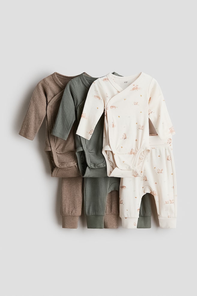 Three matching baby sets, each consisting of a long-sleeve wrap bodysuit and pants, are displayed front-facing in brown ribbed, sage green ribbed, and patterned off-white cotton jersey. The bodysuits feature side and bottom press-stud closures, while the coordinating pants have cuffed hems. - 1