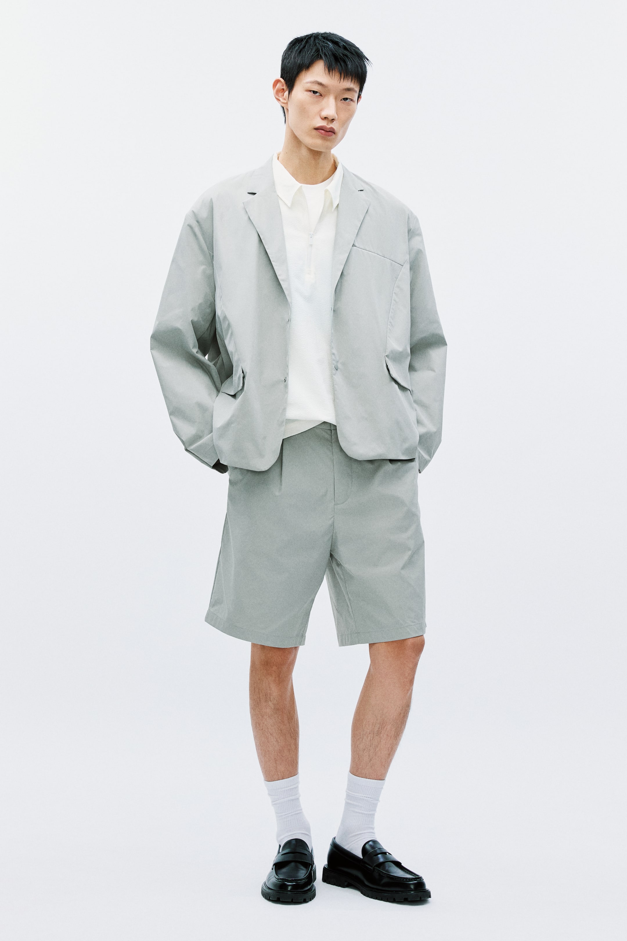 View larger image: The model is donning an open light grey blazer layered over a white collared shirt. It showcases notch lapels, a chest pocket, and front flap pockets, with press-studs prominently visible along the front.