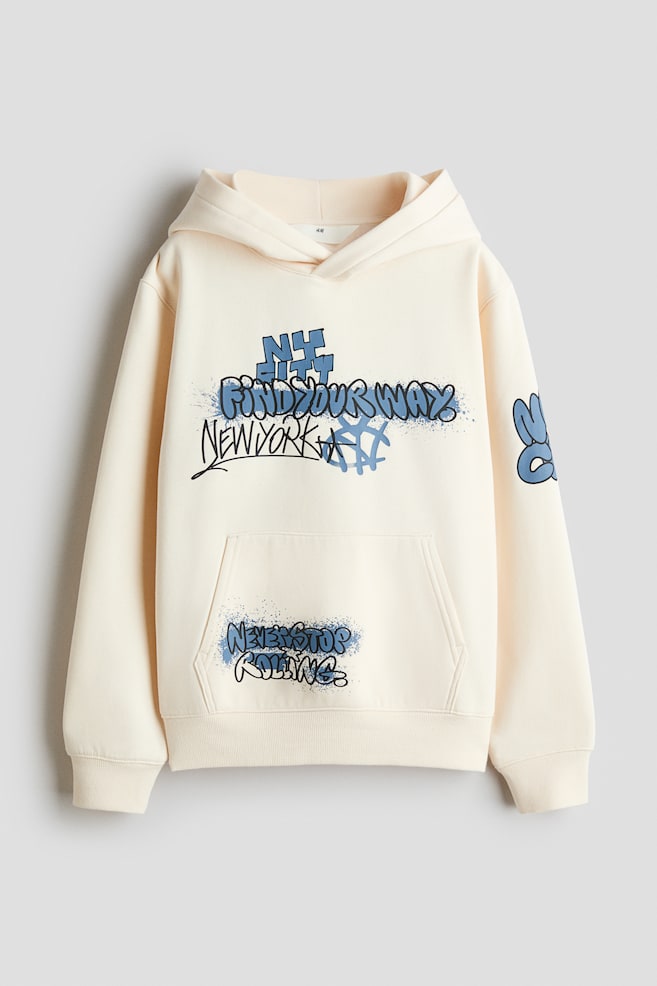 A cream-coloured hoodie is shown from the front, featuring a lined hood with a wrap front, dropped shoulders and a kangaroo pocket. The garment is decorated with blue and black graffiti-style text prints on the chest, the front pocket and the left sleeve, and is finished with ribbed cuffs and a ribbed hem. - 1